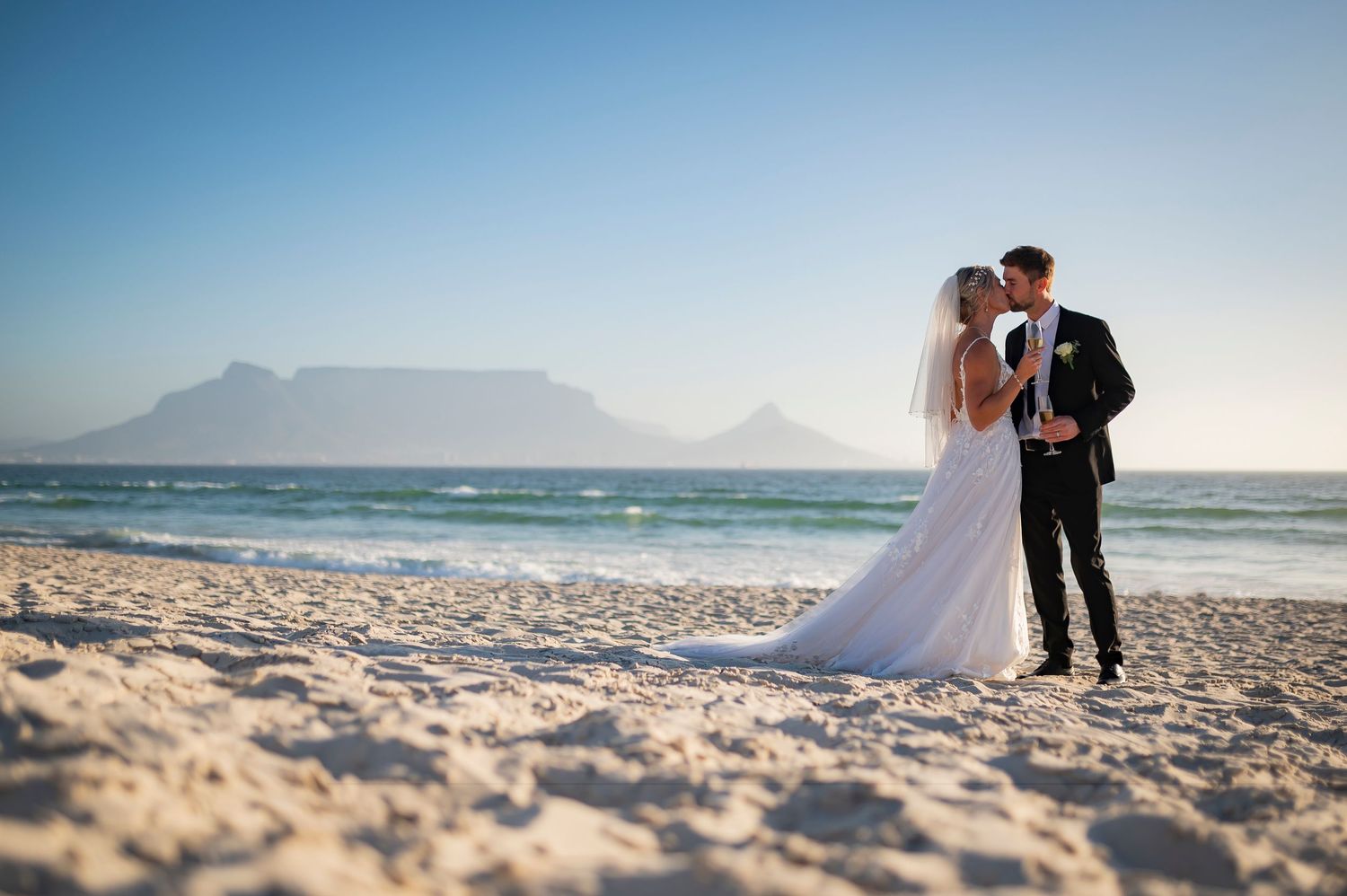 A couple in wedding attire embrace on a scenic beach with Table Mountain visible in the background at sunset.