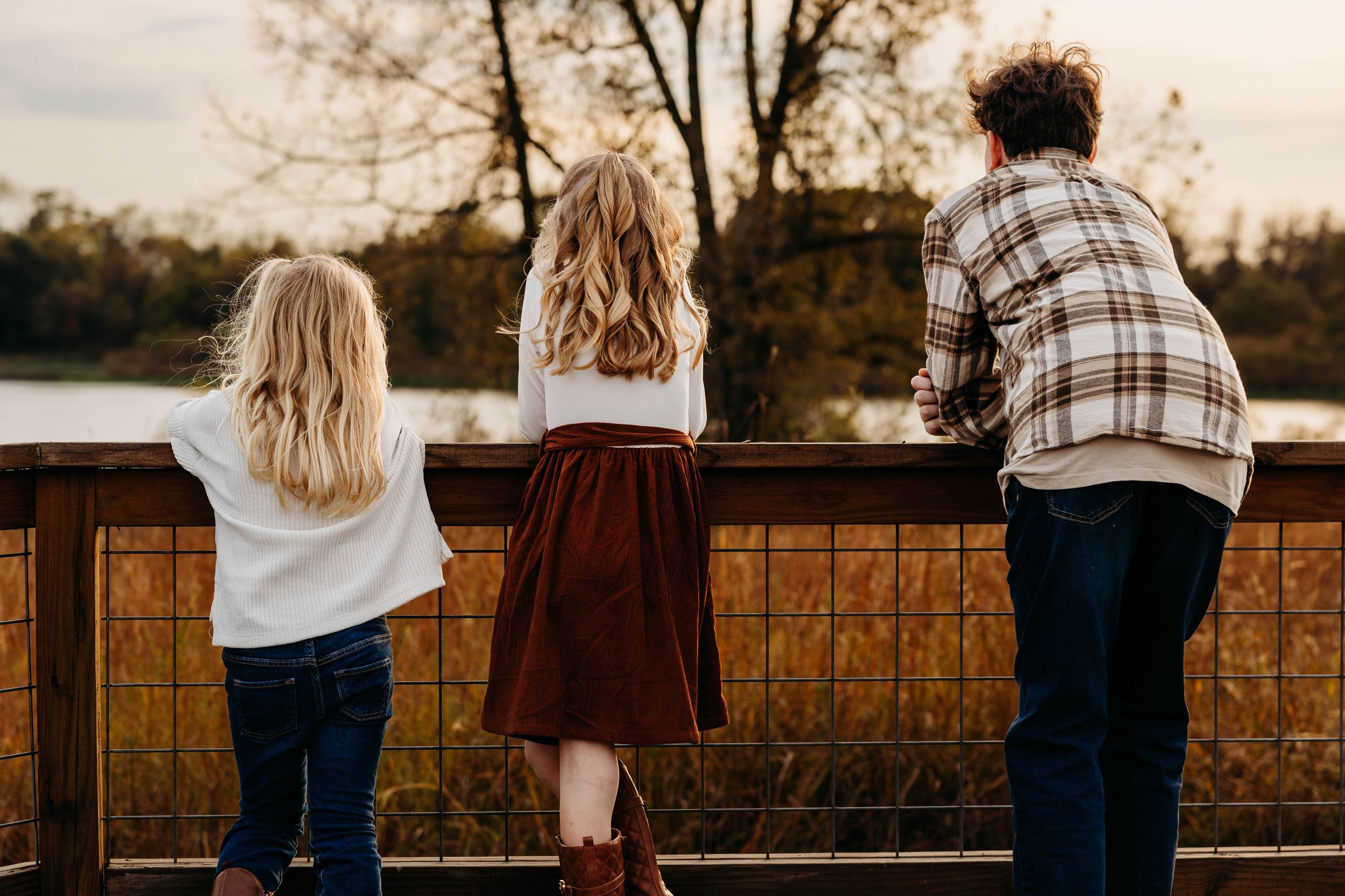 People sit on a wooden fence at sunset overlooking a scenic rural landscape in warm autumn light.