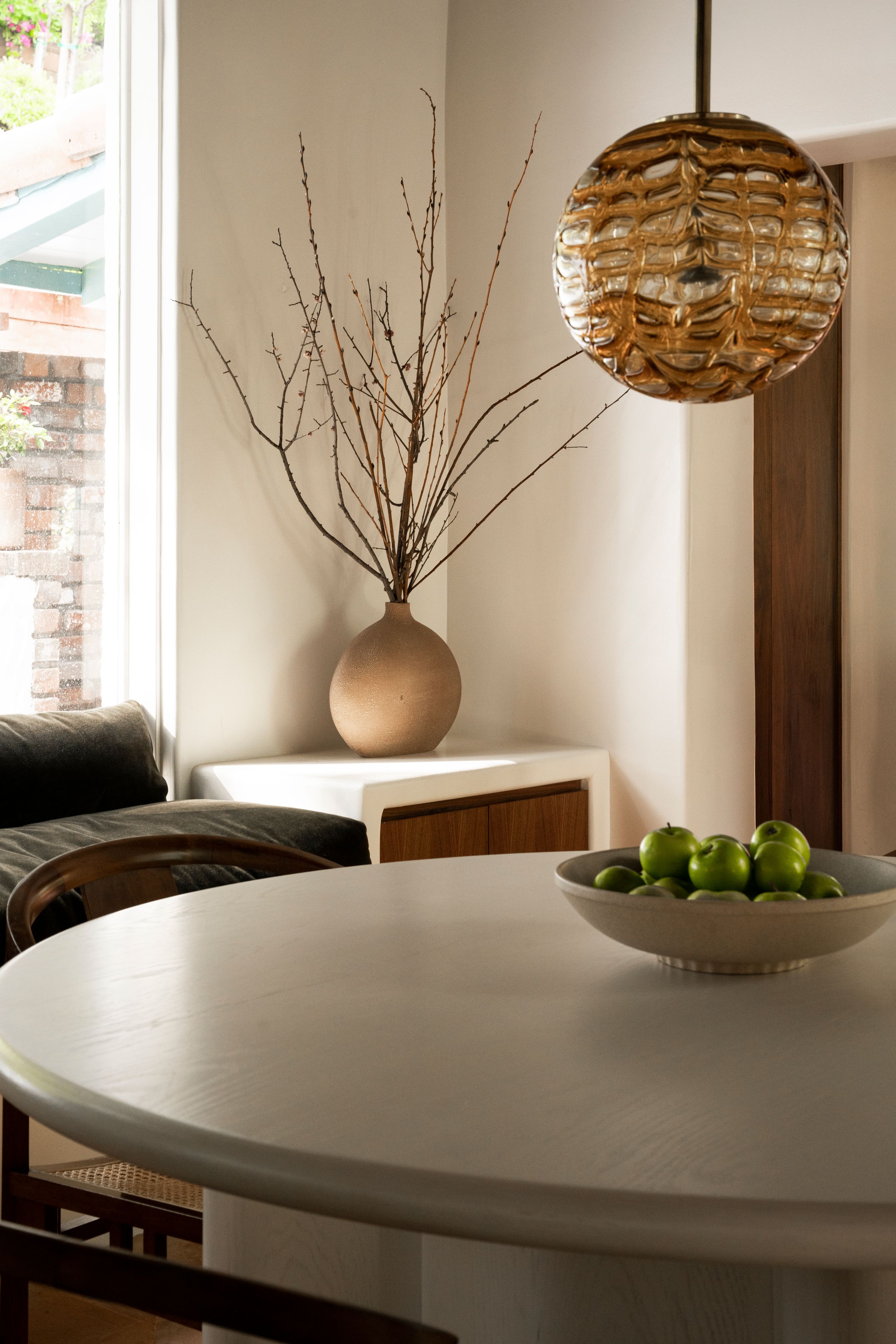 Modern dining area with round table, decorative branches in vase, and woven pendant light fixture.