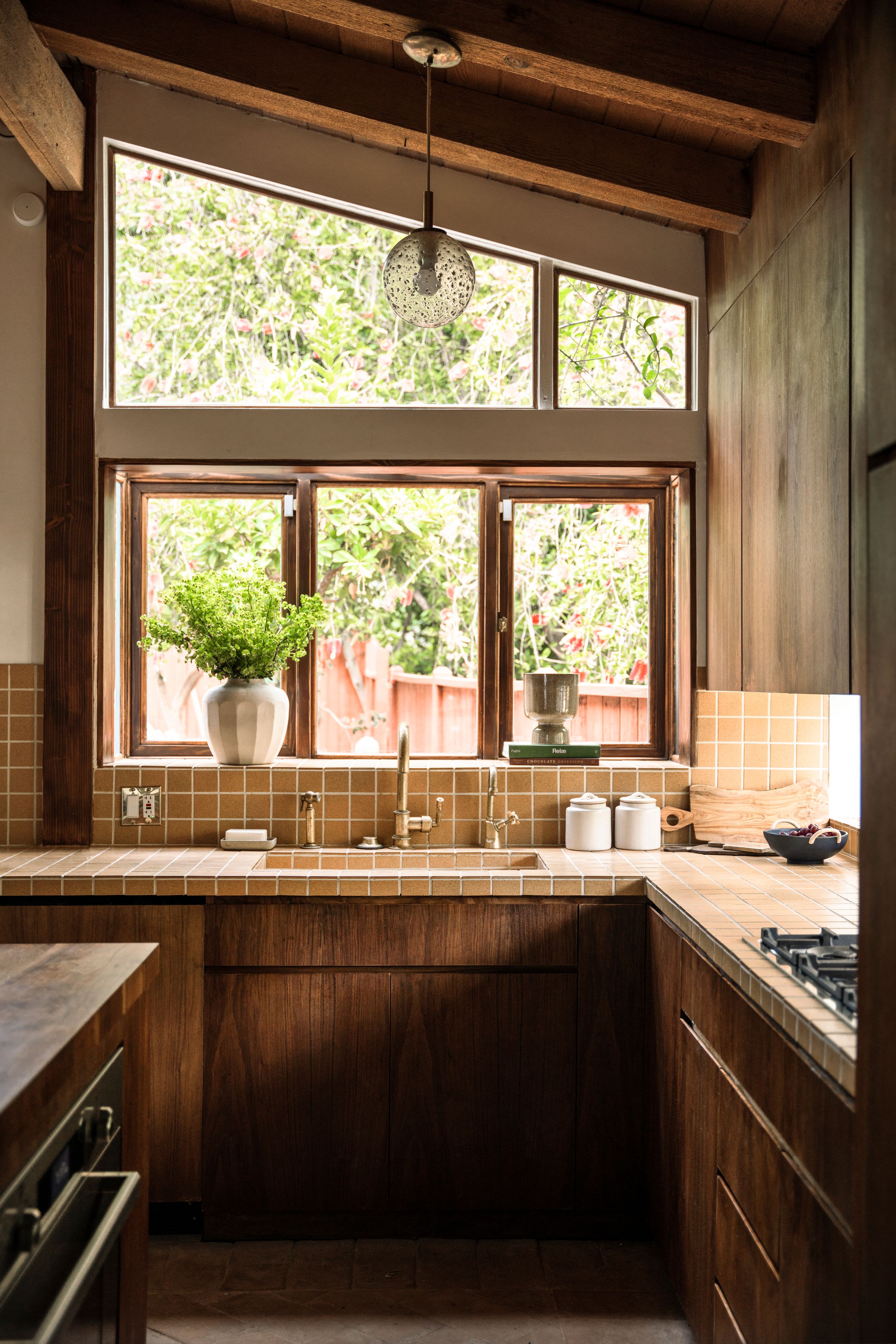 Modern kitchen with wooden cabinets, large windows, and natural light streaming through leafy garden views.