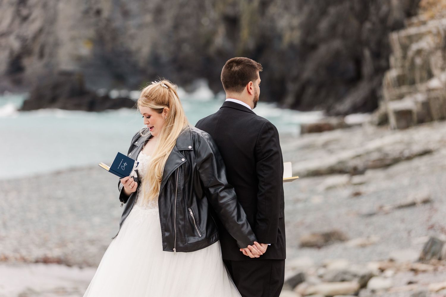 Black and white photo of couple walking along rocky coastal beach in wedding attire.