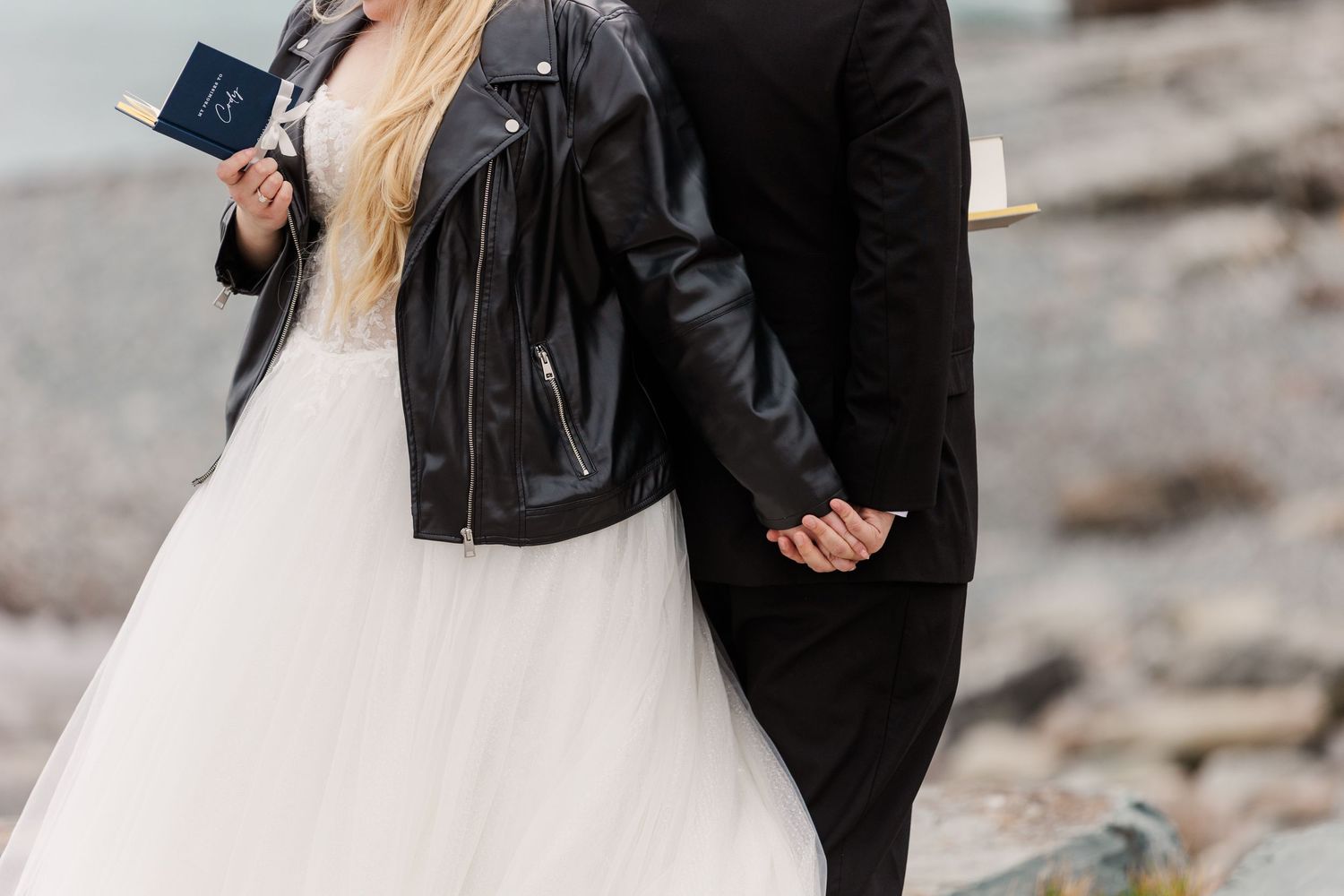 A couple in wedding attire embraces on a rocky beach, the bride wearing a flowing white dress and leather jacket.