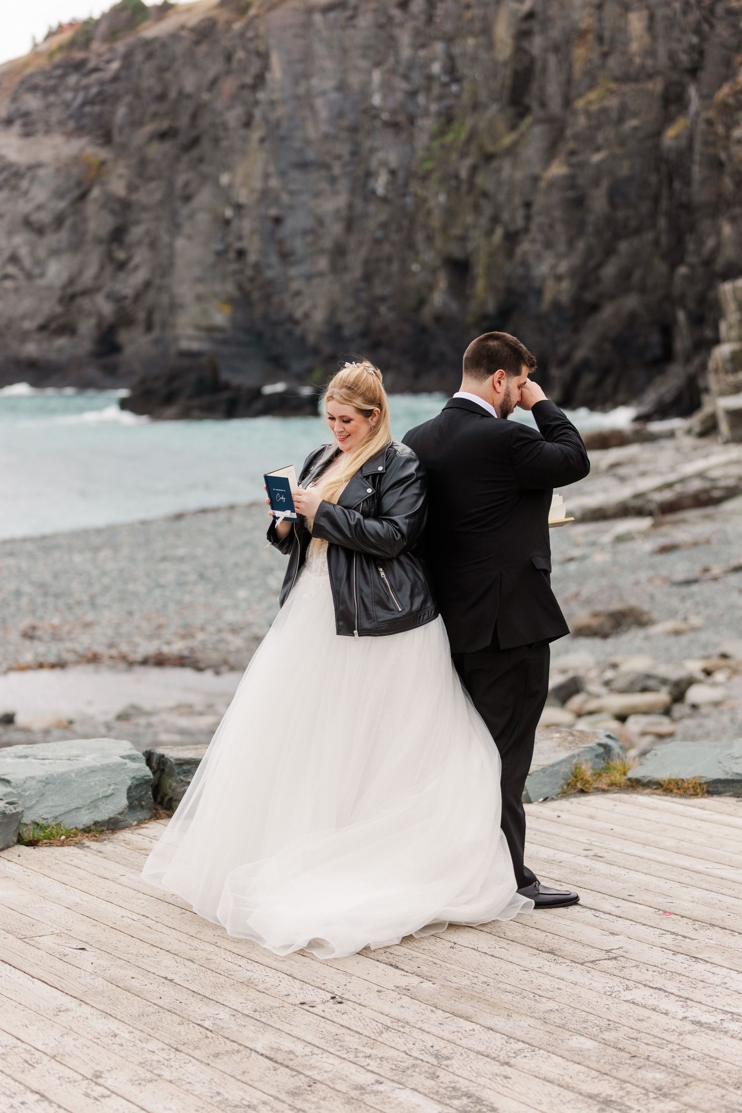 A couple in wedding attire playfully dips on a wooden boardwalk against dramatic coastal cliffs.