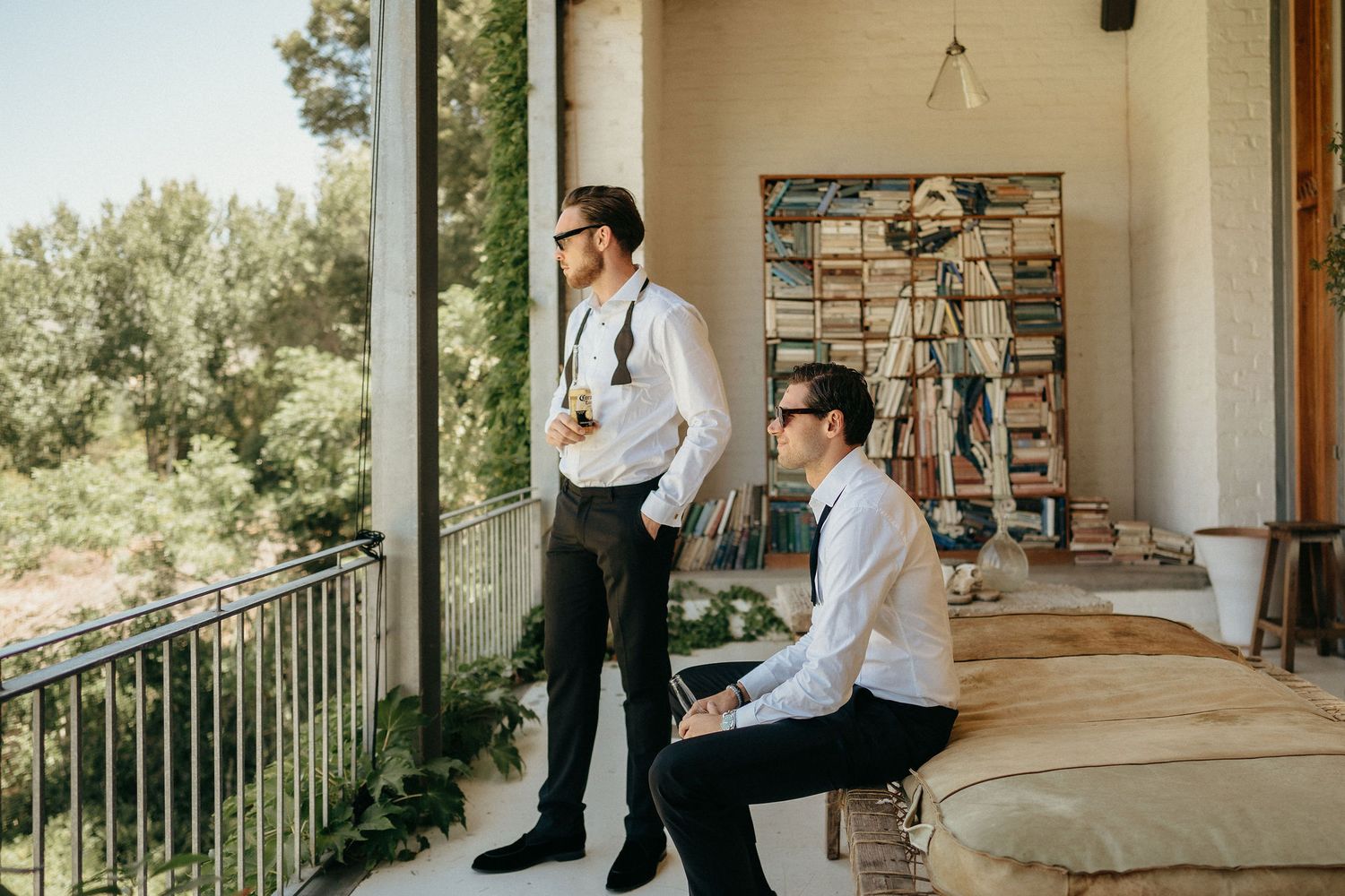 Two people in formal white shirts and dark pants stand on a sunny porch with stone walls and railing.