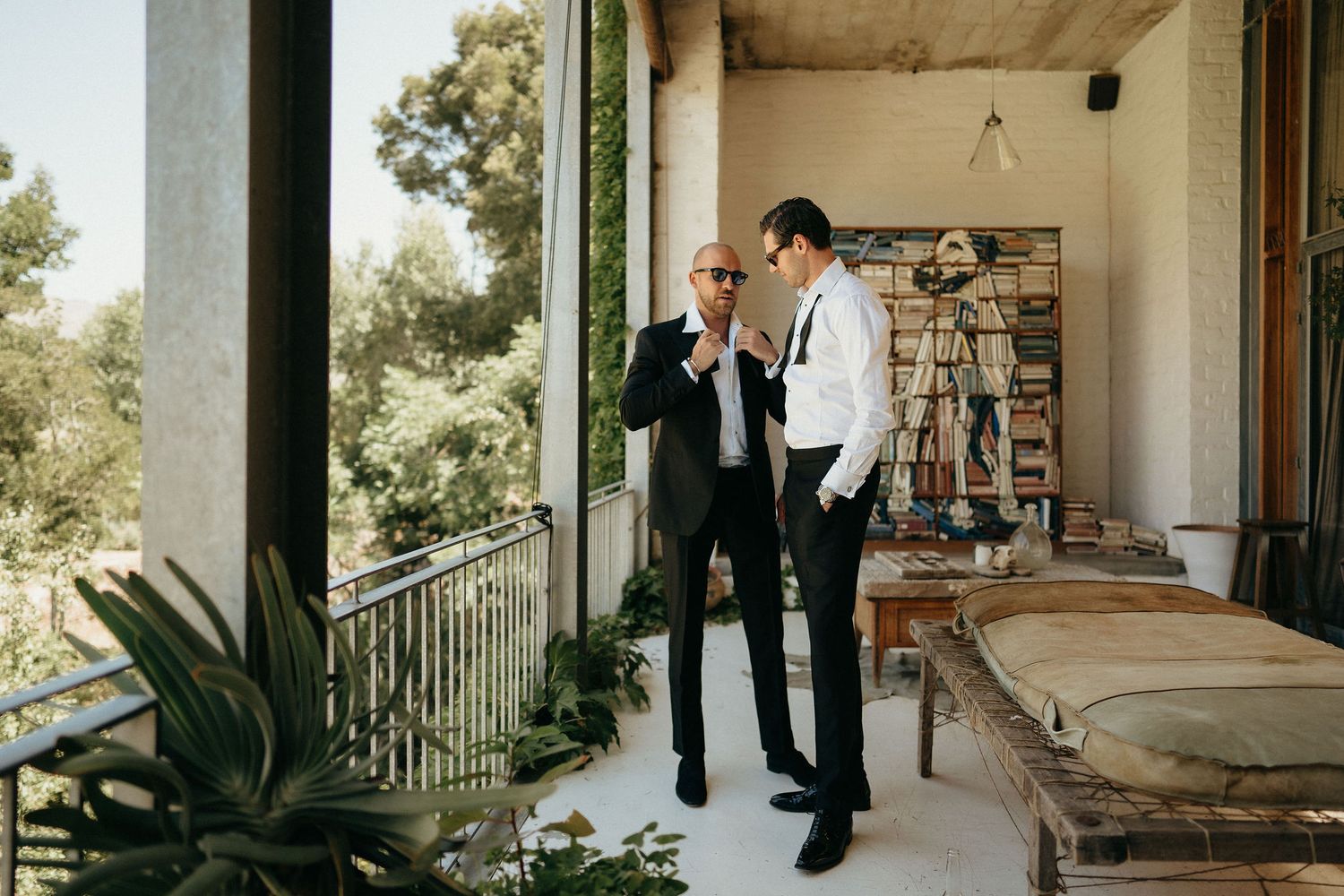 Groomsmen in formal black suits and white shirts gather on an elegant stone patio surrounded by lush greenery.