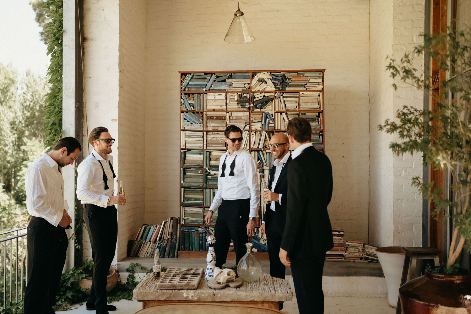 Groomsmen in formal attire gather on a covered porch with bookshelves and greenery in the background.