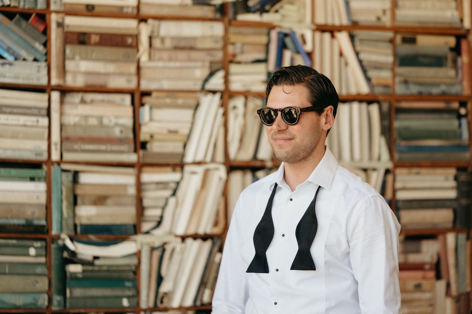 Professional wearing sunglasses and white shirt stands in front of tall bookshelves in a library or bookstore.