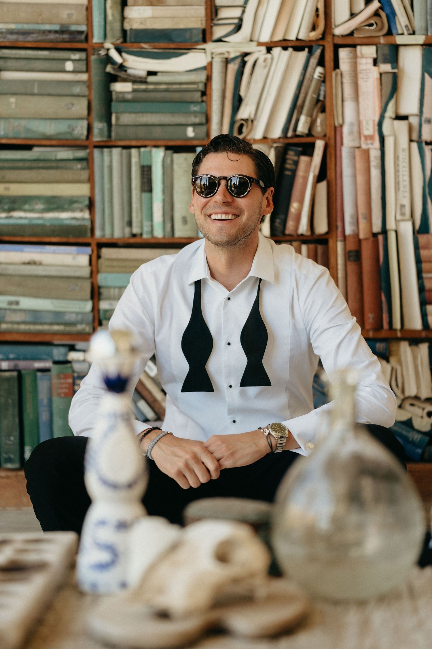 A person in a white dress shirt and sunglasses sits at a table smiling in front of bookshelves in a library setting.