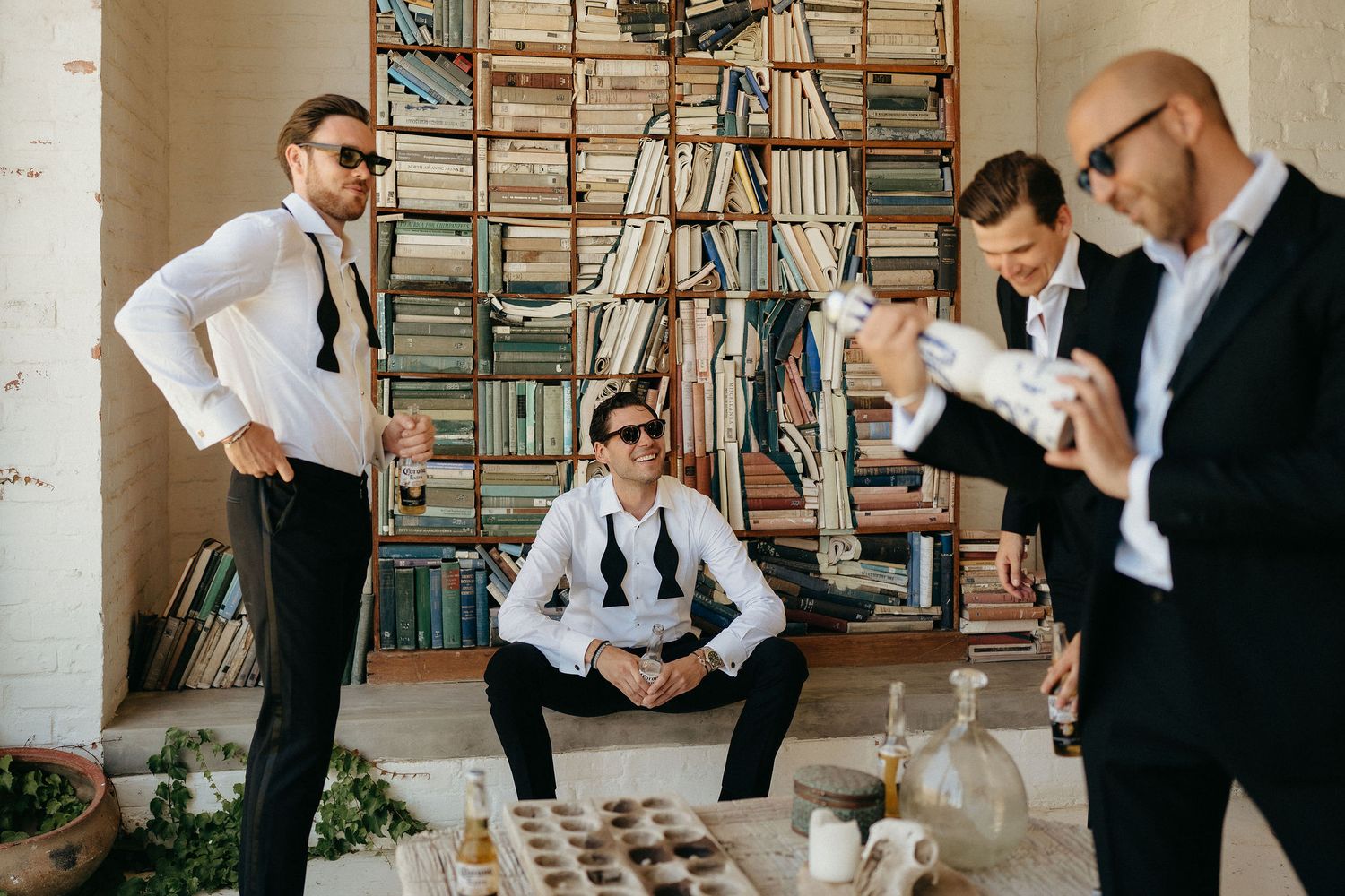 Groomsmen in formal attire gather around bookshelves while getting ready for a wedding celebration.