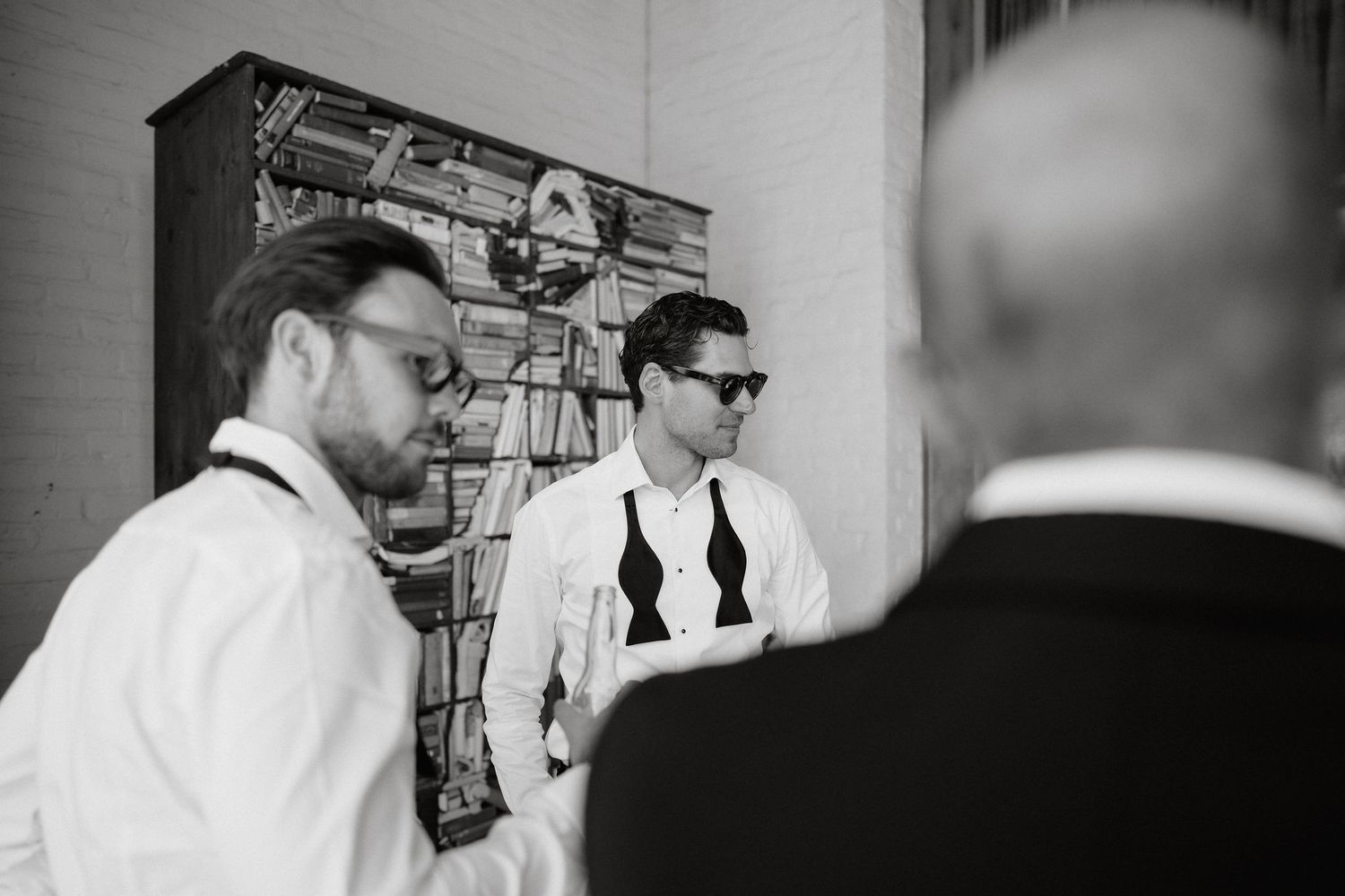 Black and white photo of wedding party members preparing in a room with vintage bookshelves.
