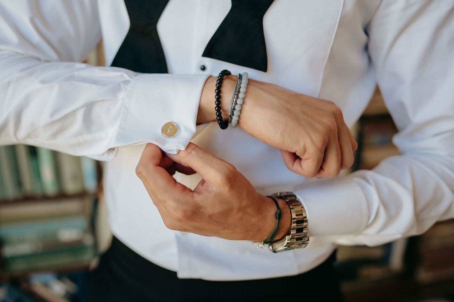 Close-up view of hands adjusting white shirt cuffs while wearing a black bowtie and beaded bracelets.