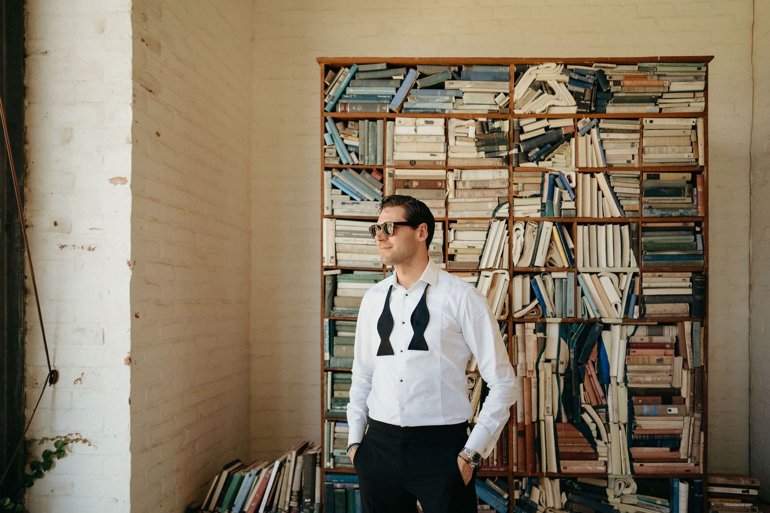Person in a white shirt and dark vest stands against a wall of stacked books and bookshelves in a bright room.