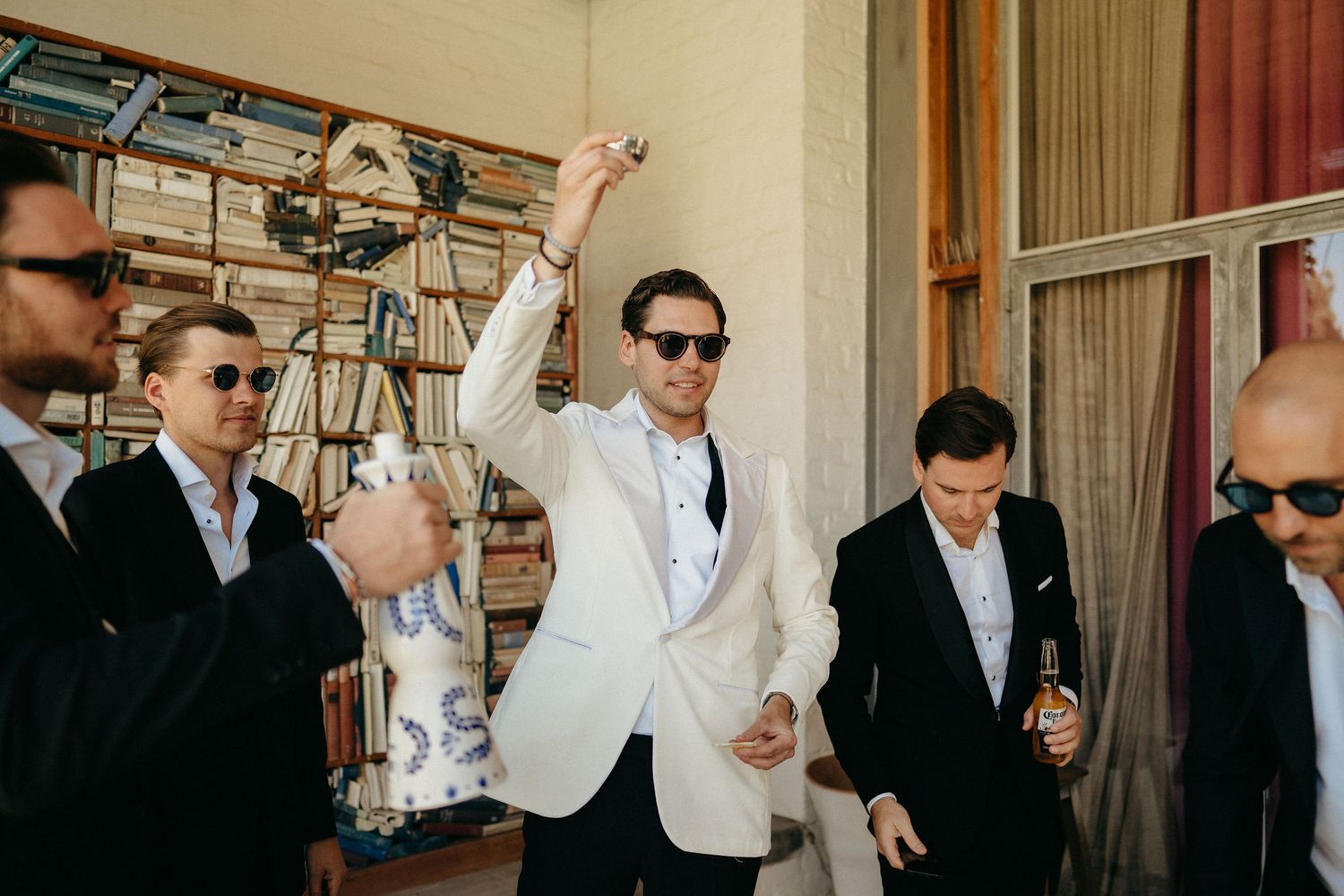 Groomsmen in dark suits and sunglasses celebrate with one member wearing a white jacket at a wedding party.