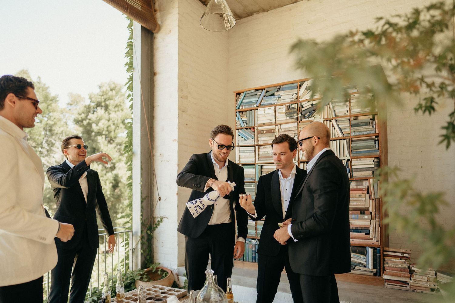 Groomsmen in black suits share laughter and drinks during a candid moment at a wedding celebration.