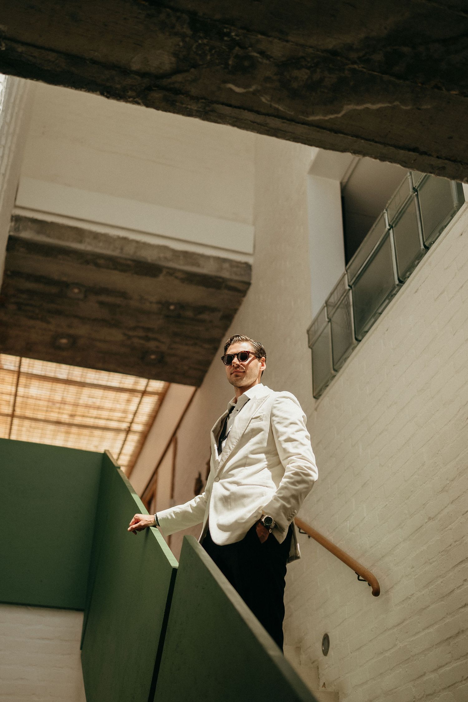 Person in white jacket and sunglasses poses dramatically in a stairwell with angular lighting and shadows.