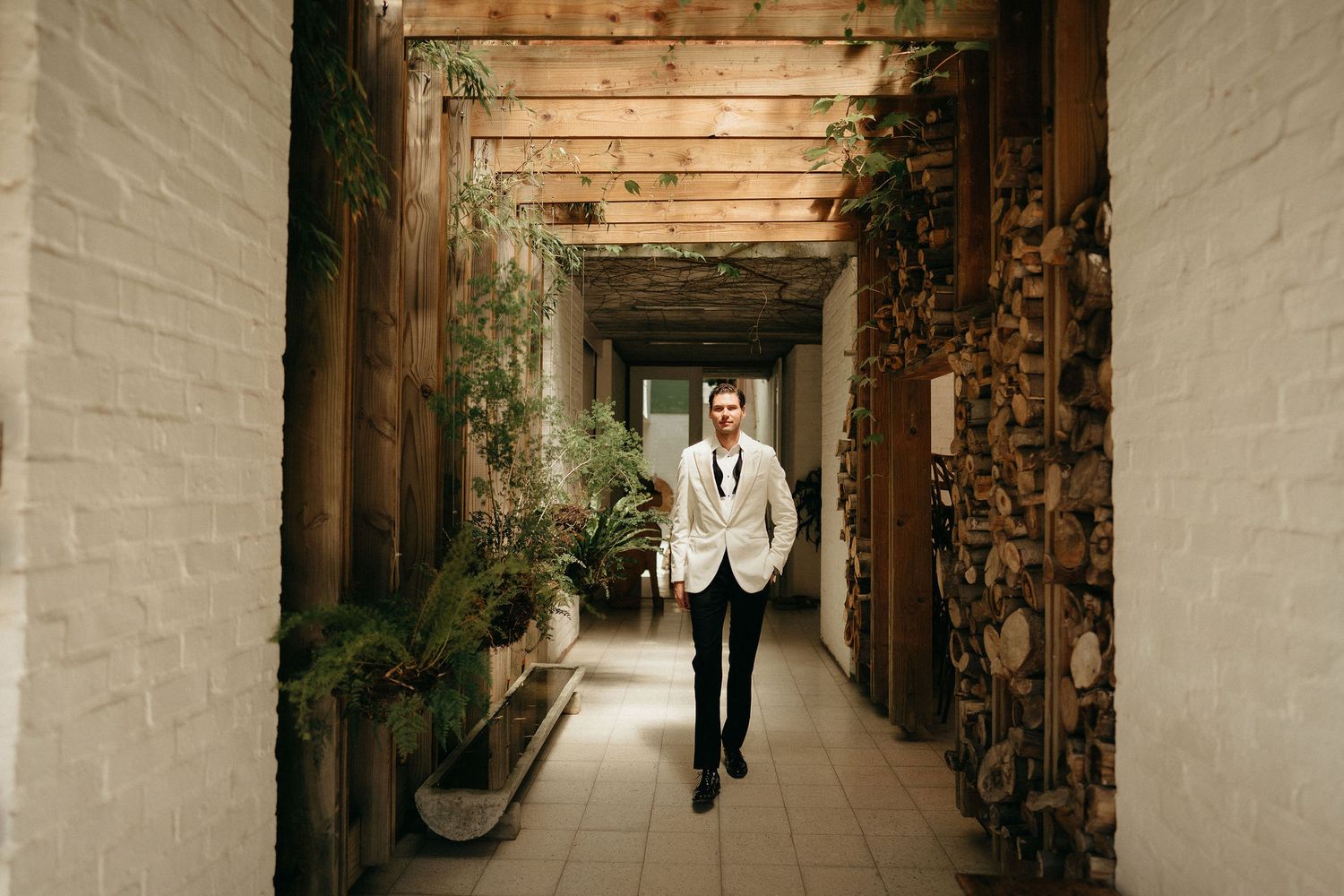 A long rustic hallway with wooden beams and white brick walls leads to a sunlit doorway at the end.