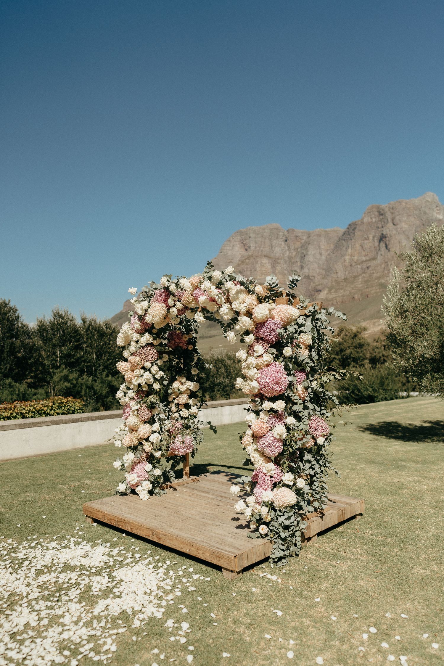 Wooden wedding arch decorated with pink and white roses against mountain backdrop with scattered flower petals on grass.