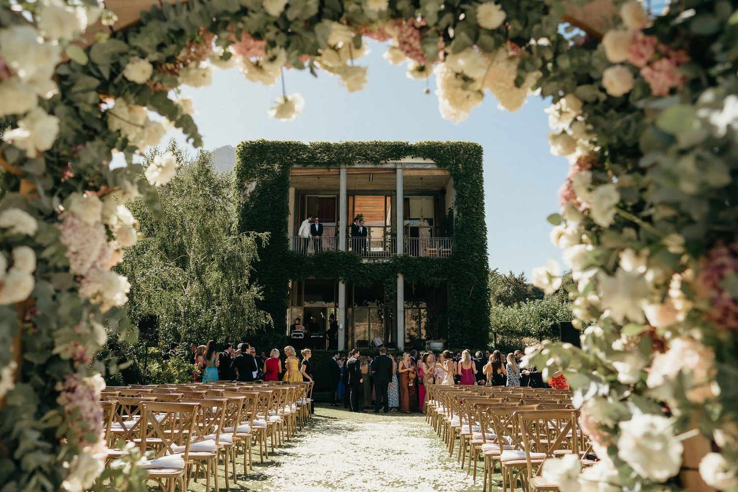 Elegant outdoor wedding ceremony setup with floral arch and white chairs facing an ivy-covered historic mansion.