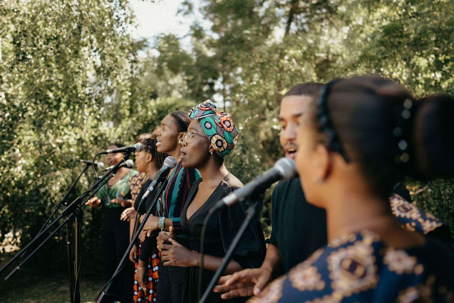 A group of people stand outdoors against a sunlit natural backdrop with trees and foliage.