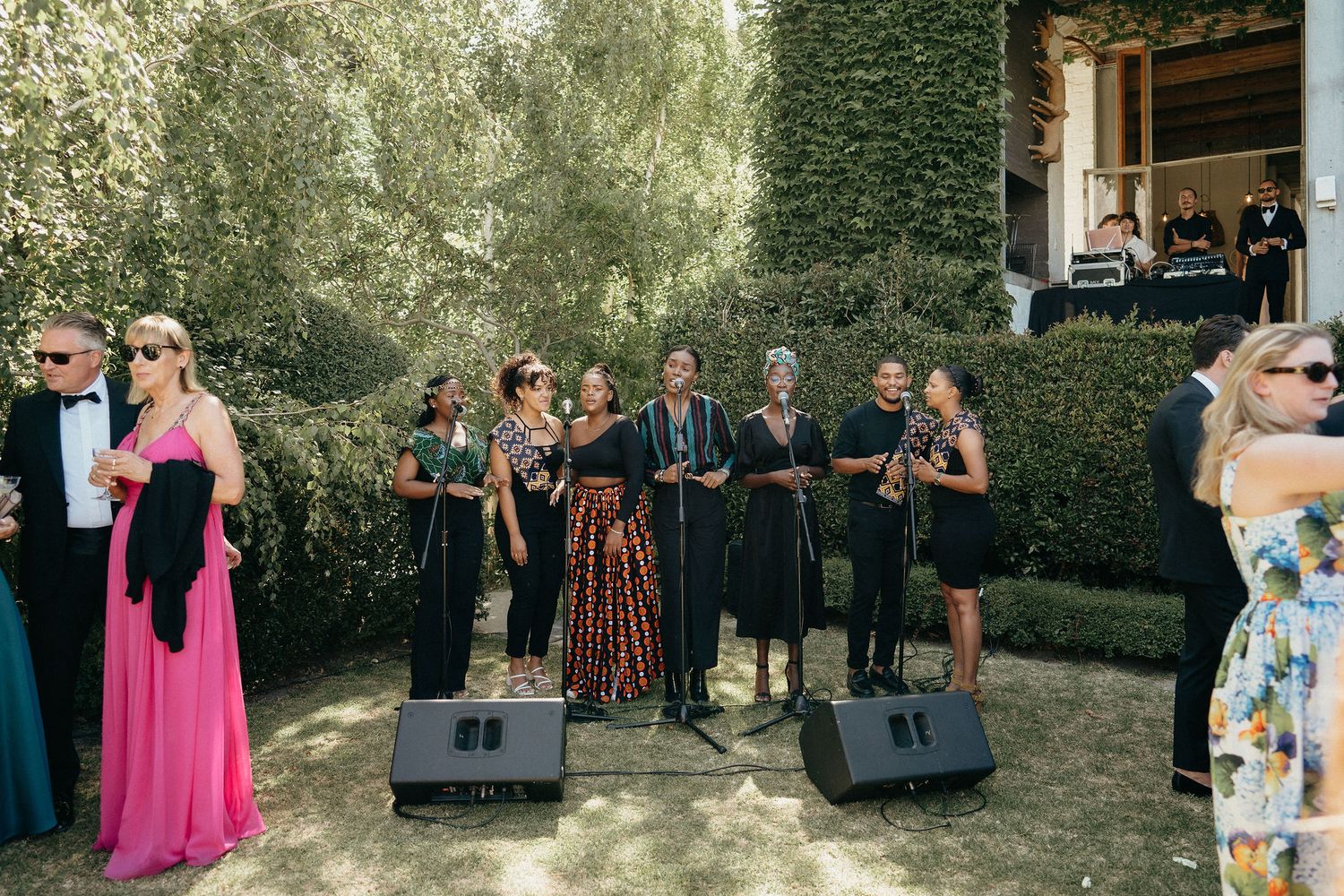 Wedding guests gather outdoors in a garden setting with lush green foliage and hedges in the background.