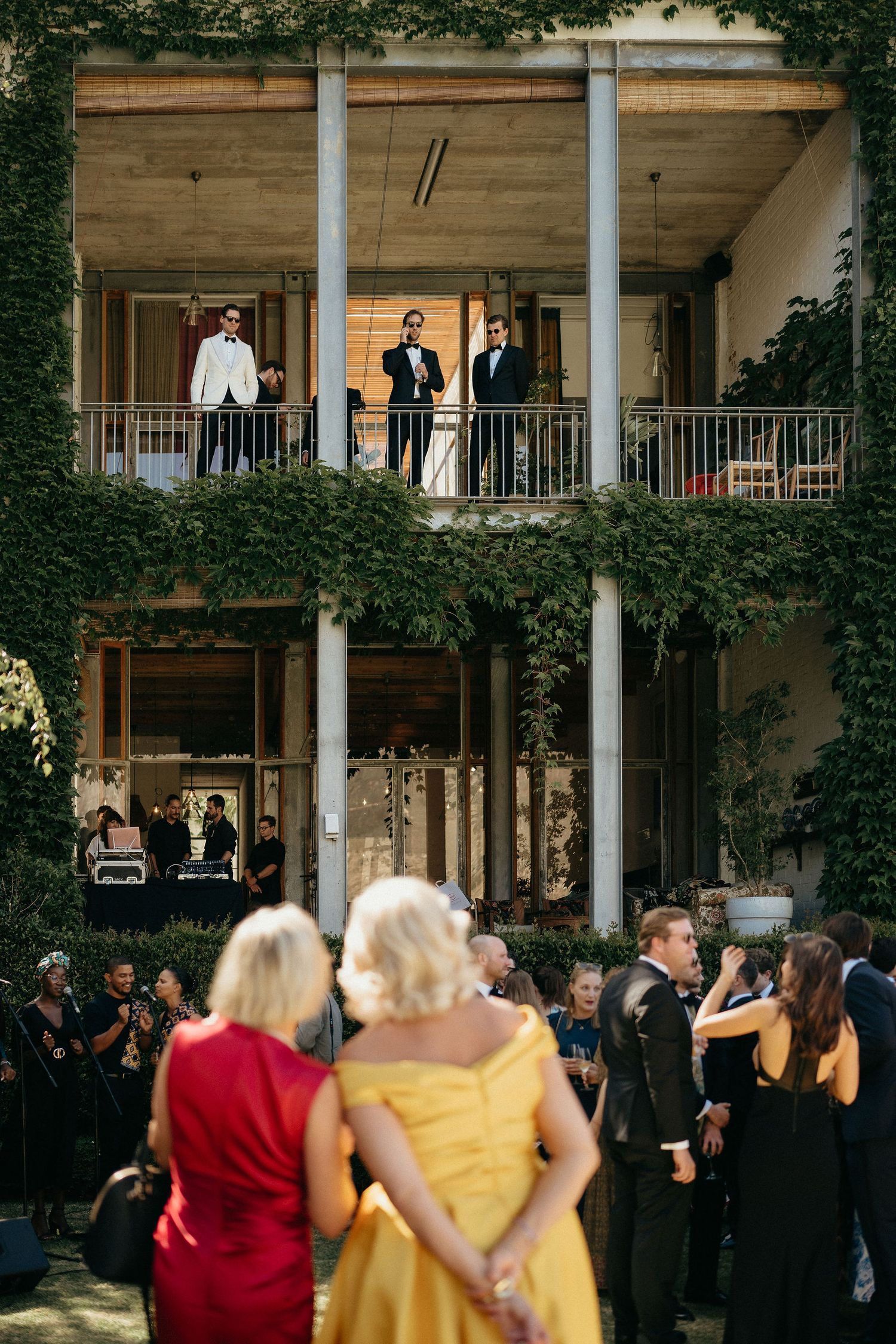 Wedding guests gather on a two-story veranda with ivy-covered columns during an outdoor reception.