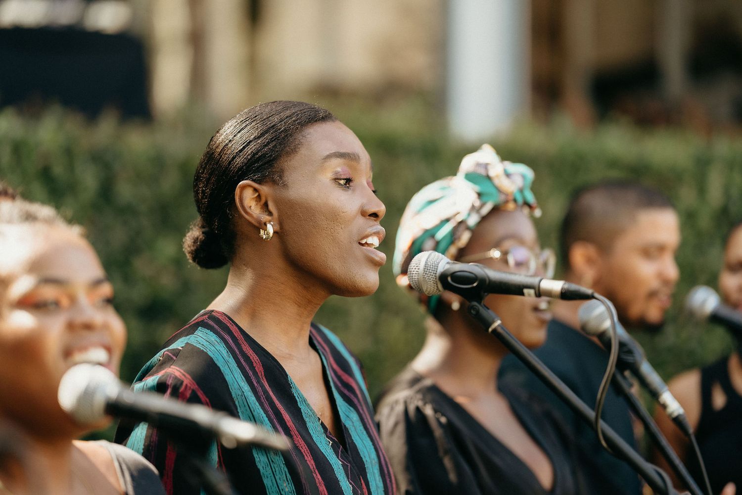 A group of performers singing together in a choir while wearing dark colored robes during an outdoor performance.