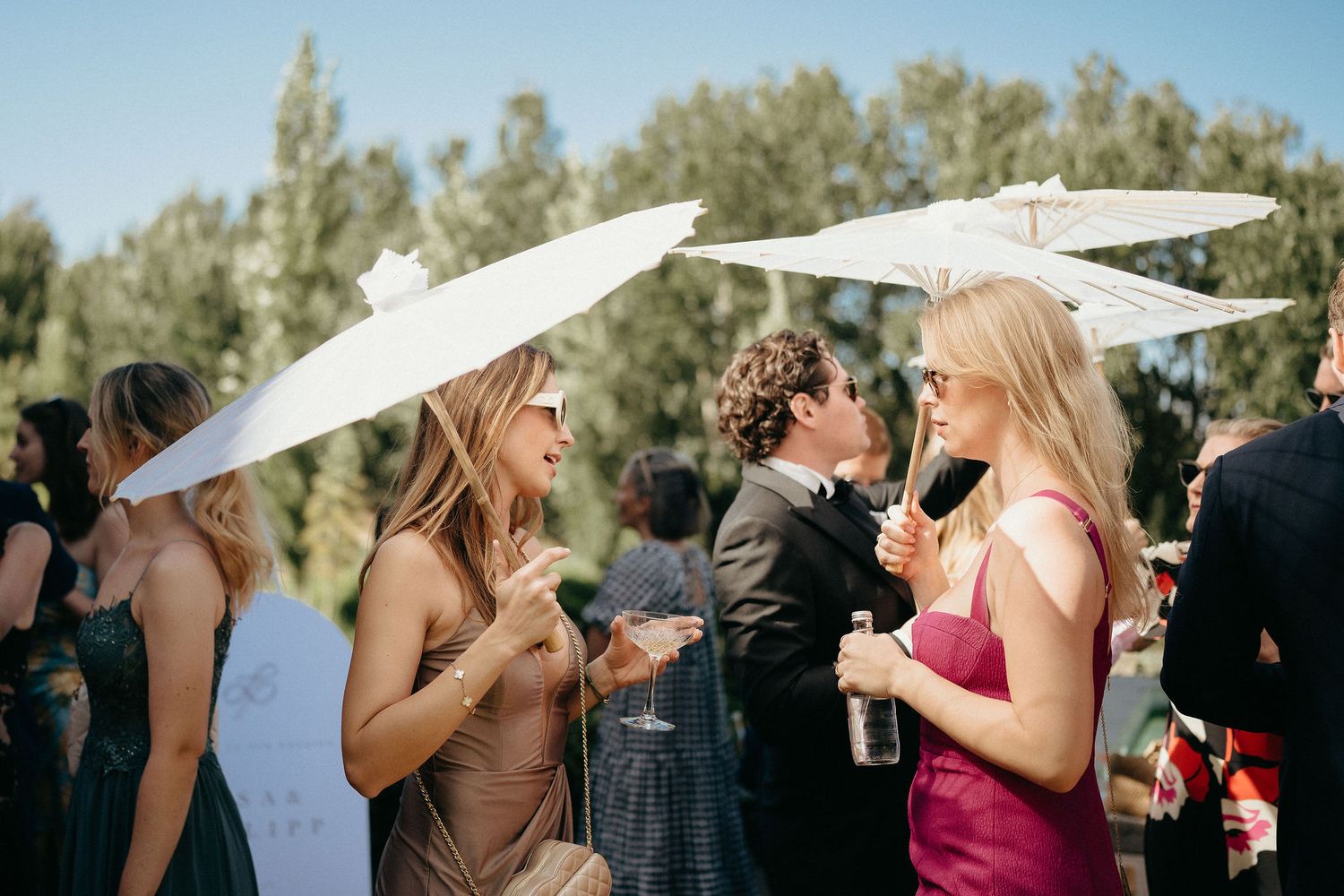 Group of wedding guests laughing and embracing while holding white parasols outdoors during sunny celebration.