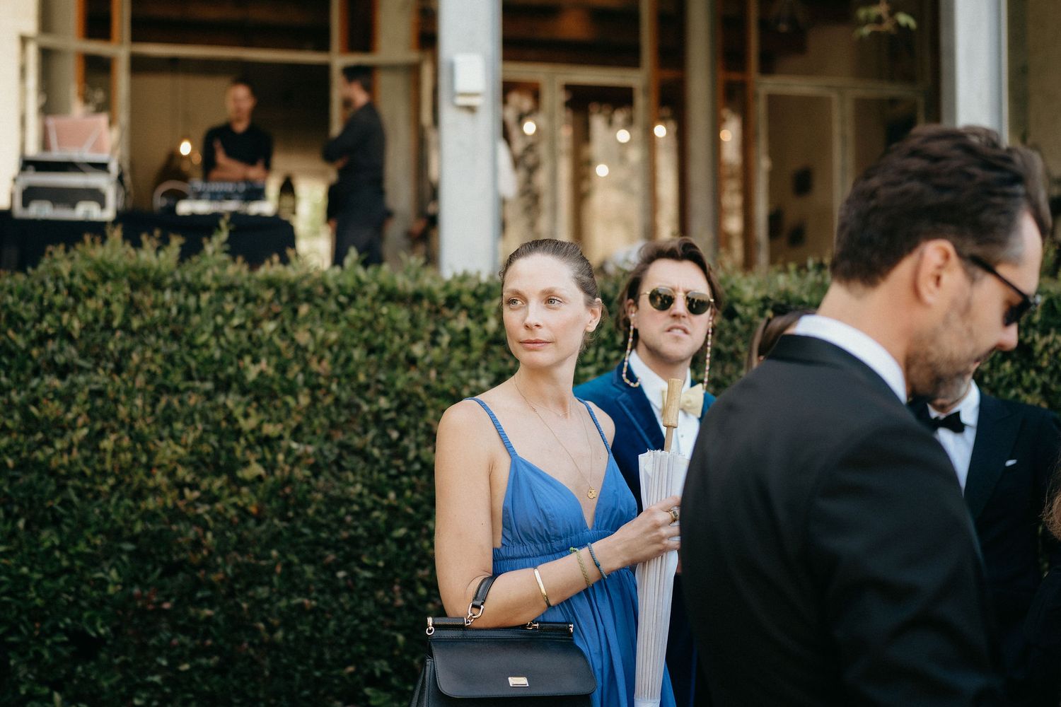 Wedding guests in formal attire gather at an outdoor reception venue with lush greenery in the background.