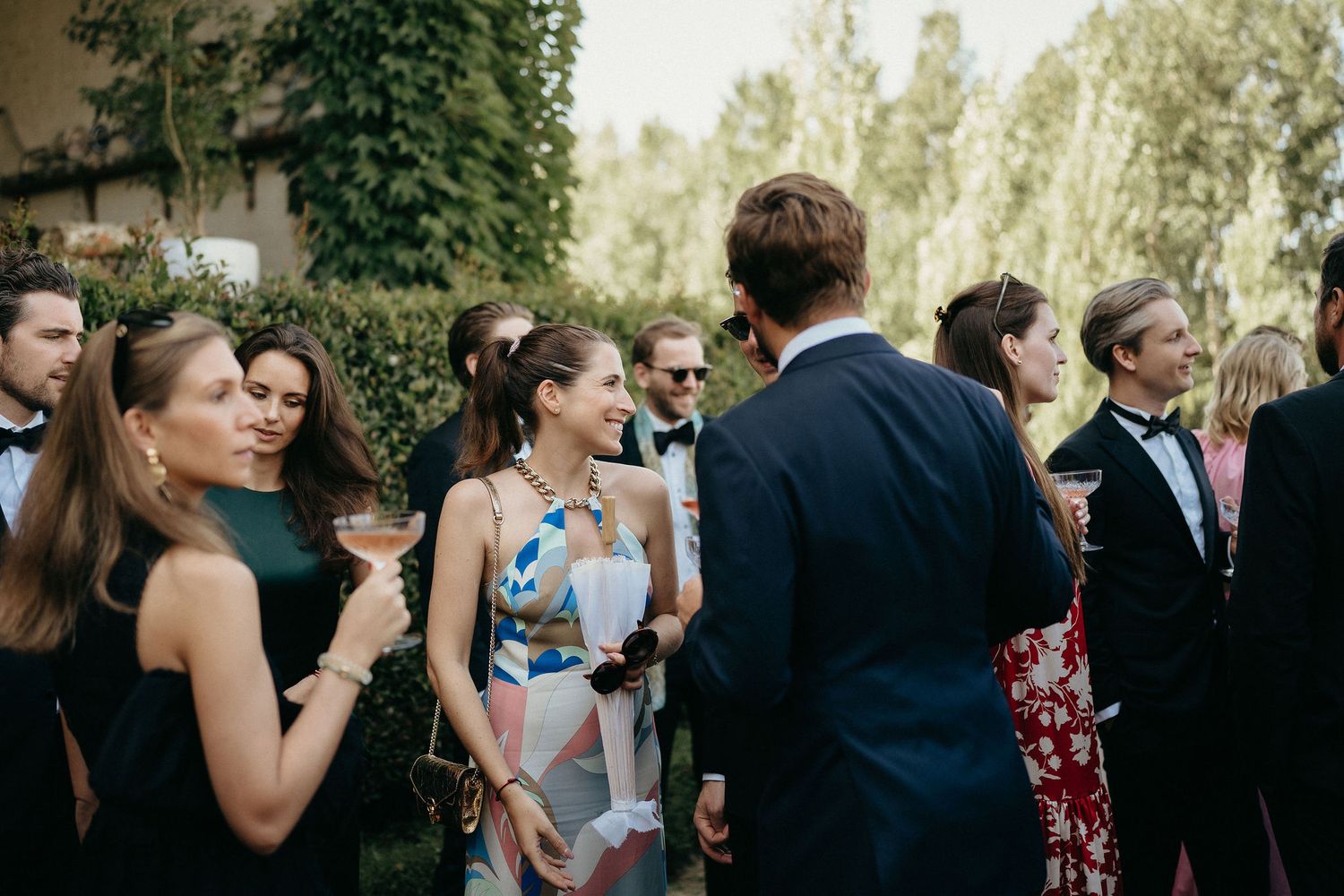 Wedding guests gather outdoors for a celebration in a garden setting with trees in the background.