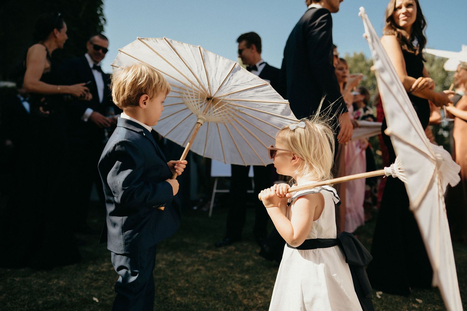 Wedding guests holding white parasols dance and celebrate at an outdoor reception during golden hour.