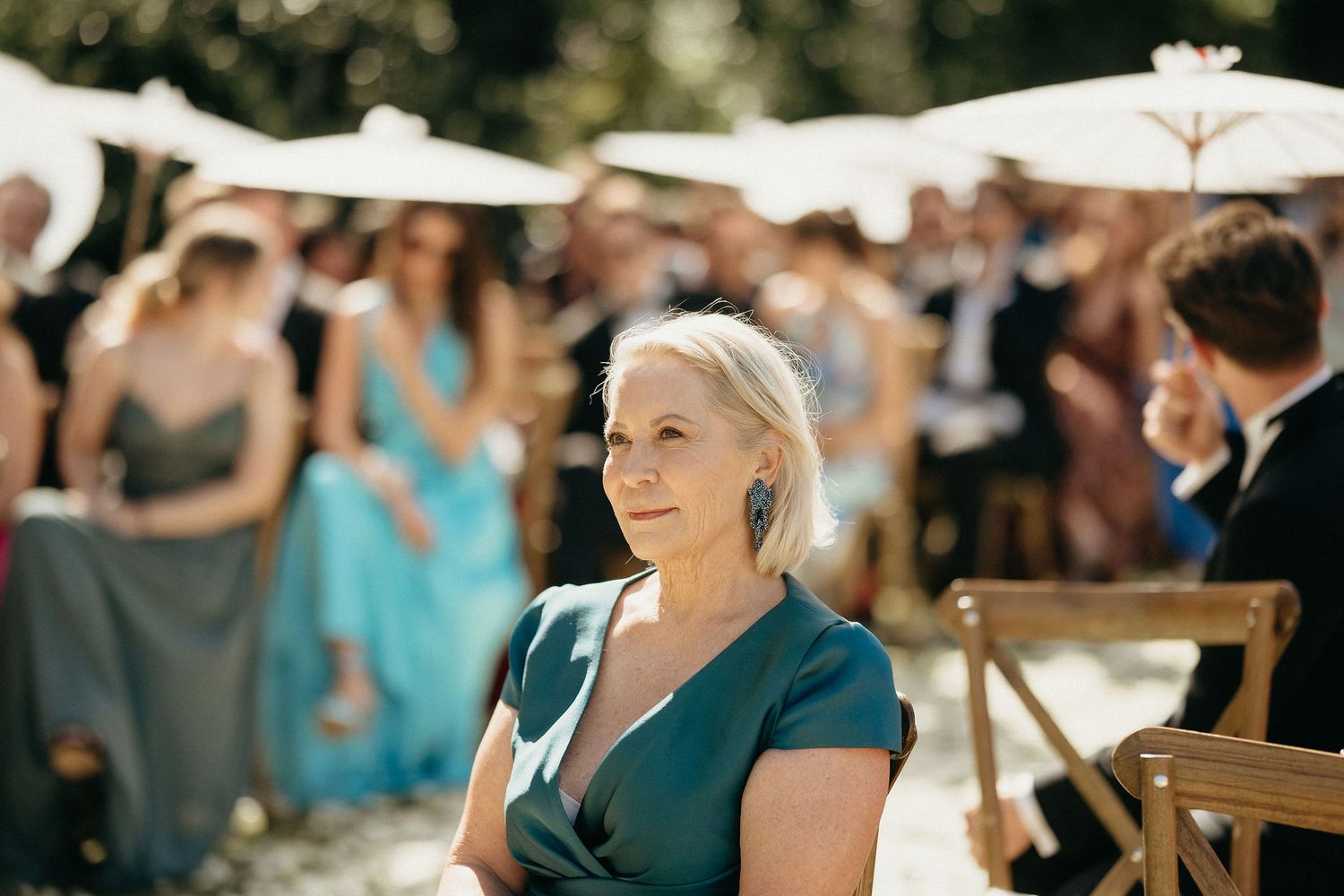 Guests seated at an outdoor wedding ceremony venue with wooden chairs and natural decor in sunlit surroundings.