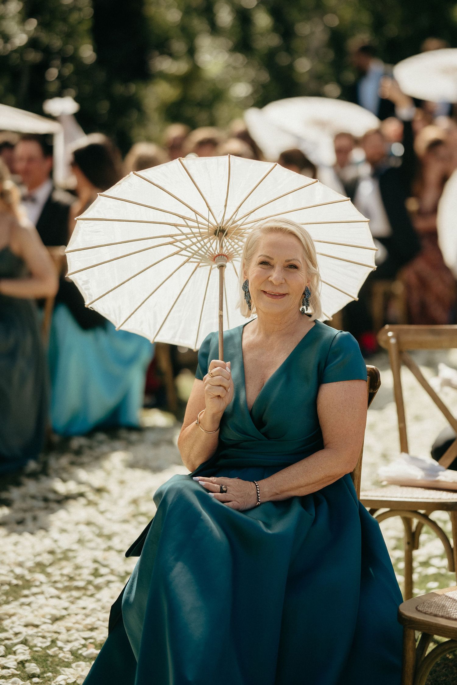 A woman in a teal dress holds a white parasol while seated at an outdoor wedding ceremony.