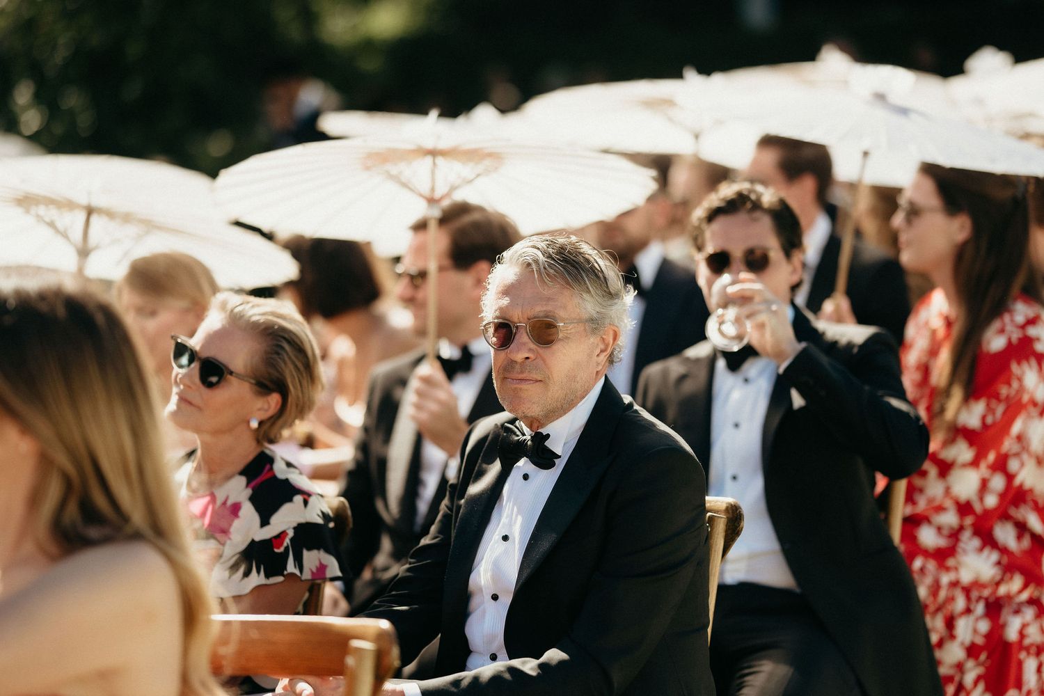 Wedding guests seated outdoors beneath white umbrellas during a sunny afternoon ceremony.