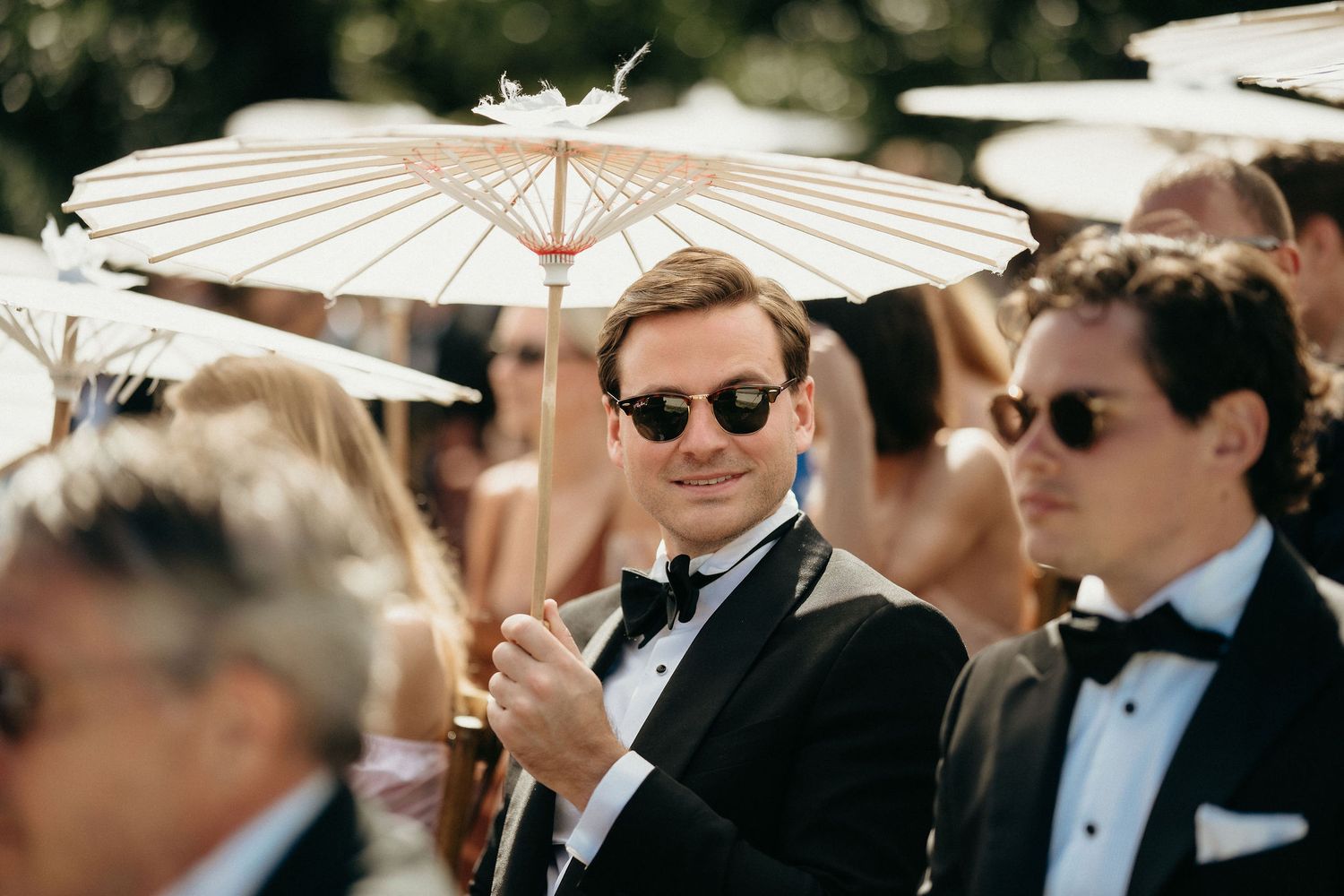 Wedding guests in formal black tie attire holding white paper parasols at an outdoor celebration.