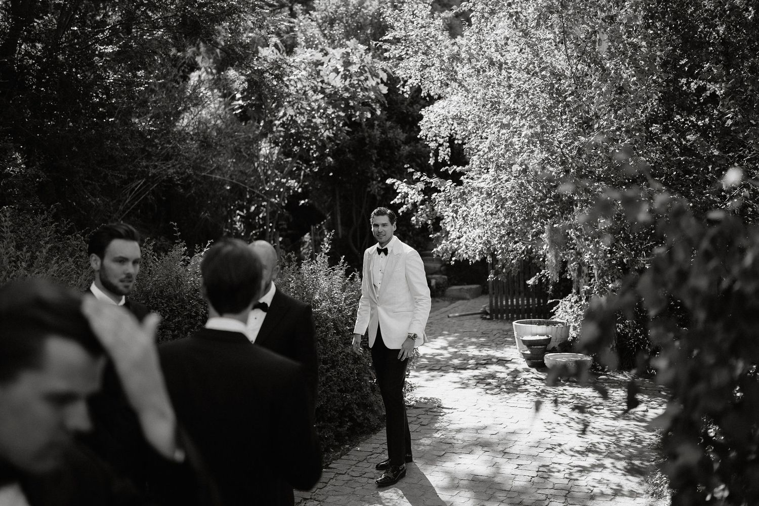 A couple walks together through a sunlit garden path surrounded by blooming flowers and lush greenery.