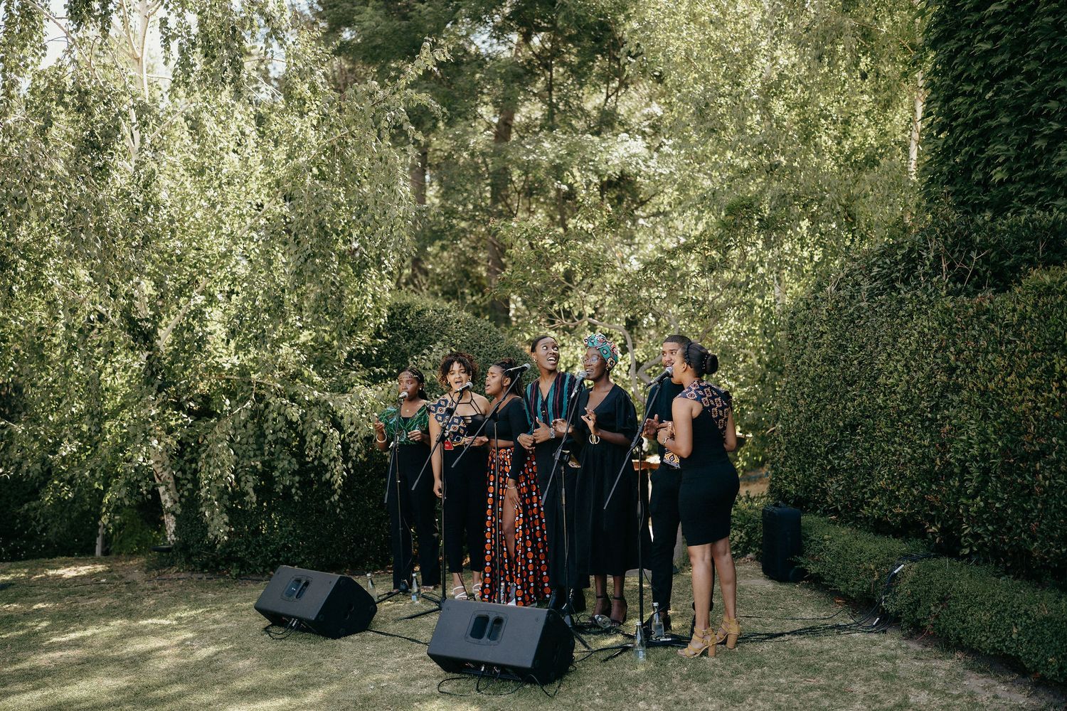 A group gathers under lush green trees in a garden setting during an outdoor event.