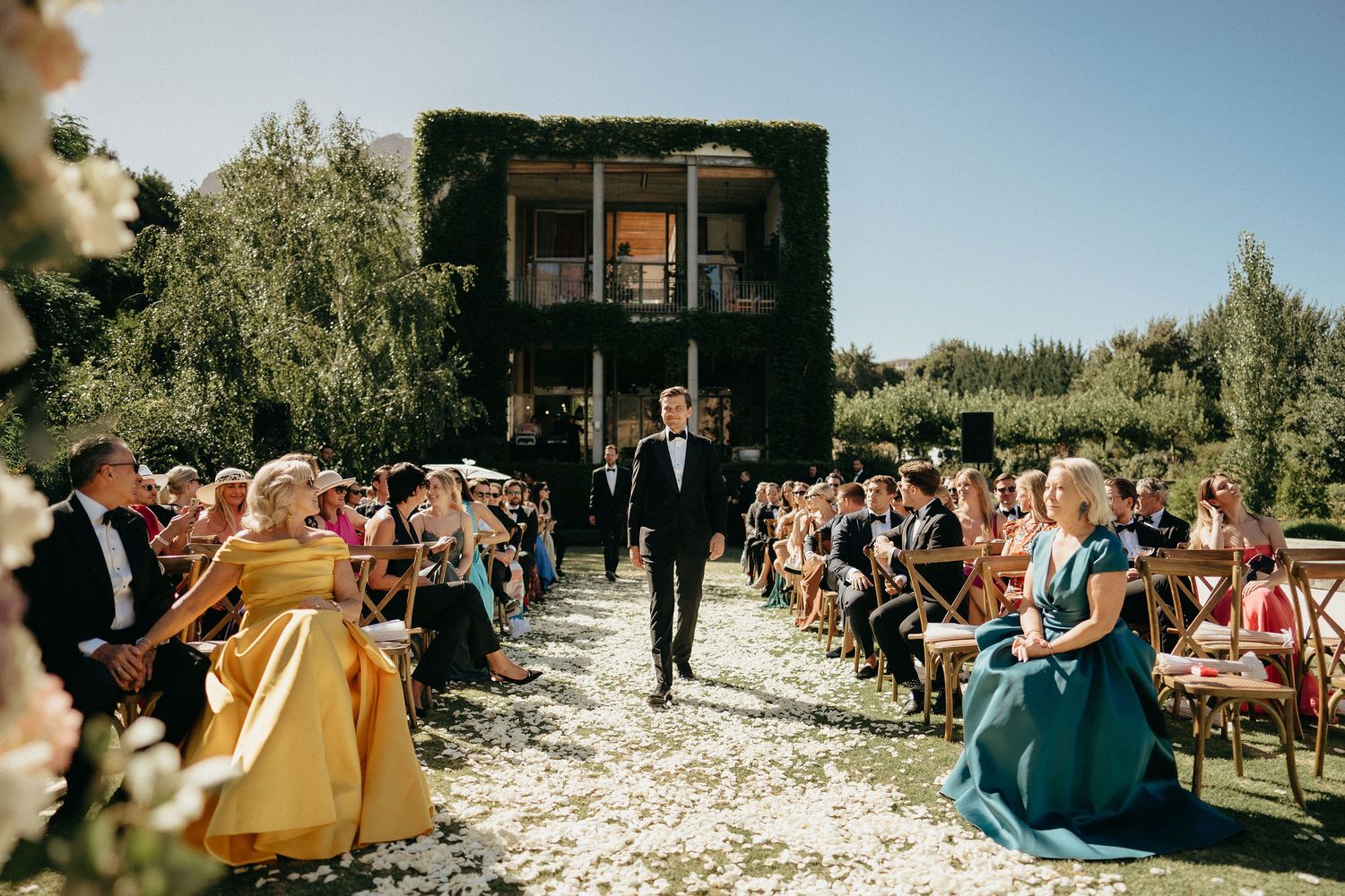 Outdoor wedding ceremony setup with white chairs and floral arrangements along a garden aisle.