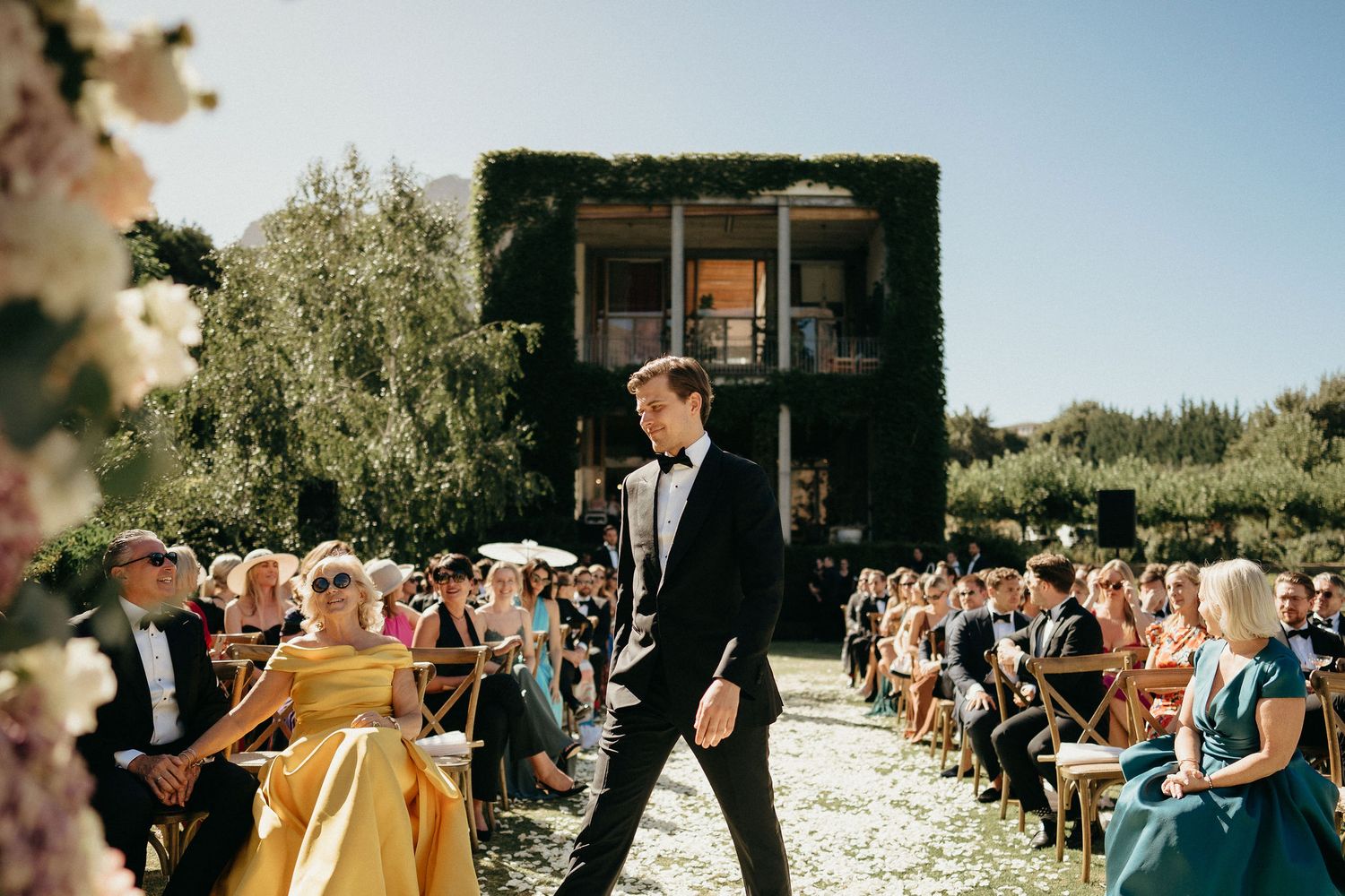 Outdoor wedding ceremony setup with white chairs and floral arrangements along a garden aisle.