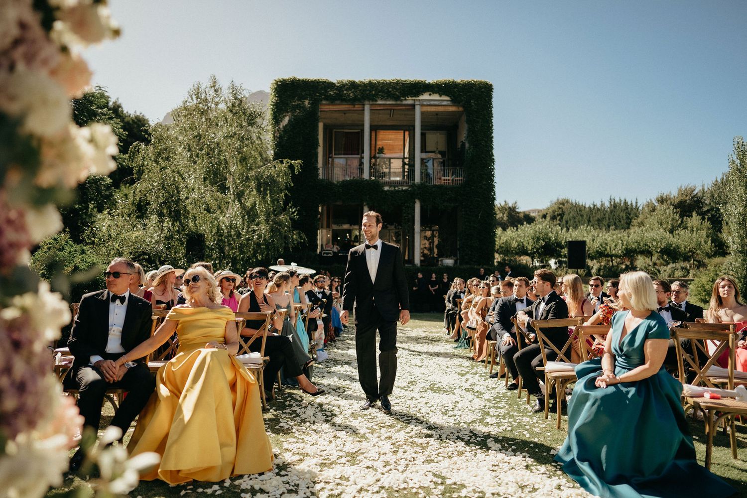 Outdoor wedding ceremony guests seated in rows along a garden path with scattered white flower petals.