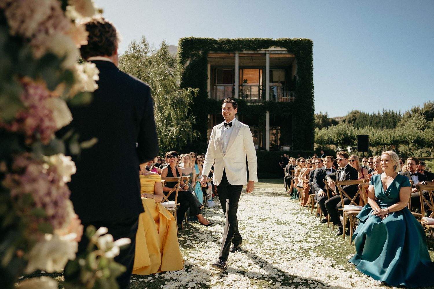 A wedding ceremony takes place outdoors with white flower petals scattered along the aisle and guests seated on both sides.