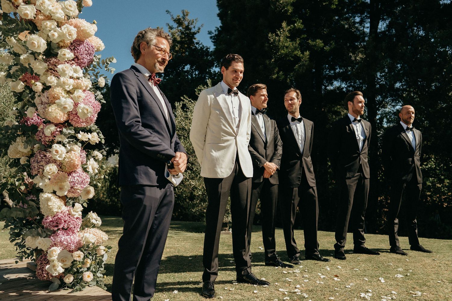 Wedding party members in dark suits stand together outdoors near elegant floral arrangements in pink and white.