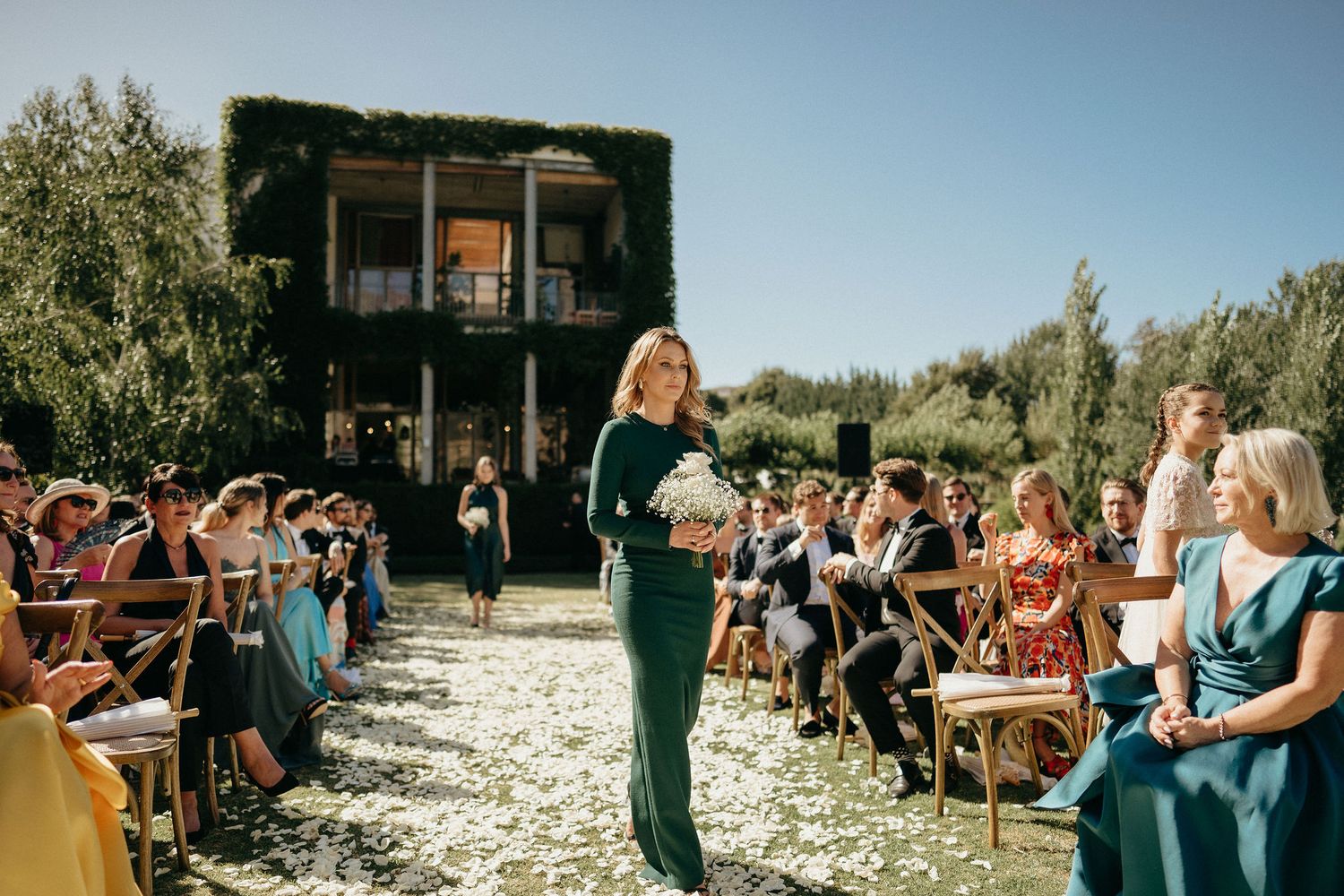 Outdoor beach wedding ceremony with guests seated in white chairs facing a wooden arch on a sunny day.