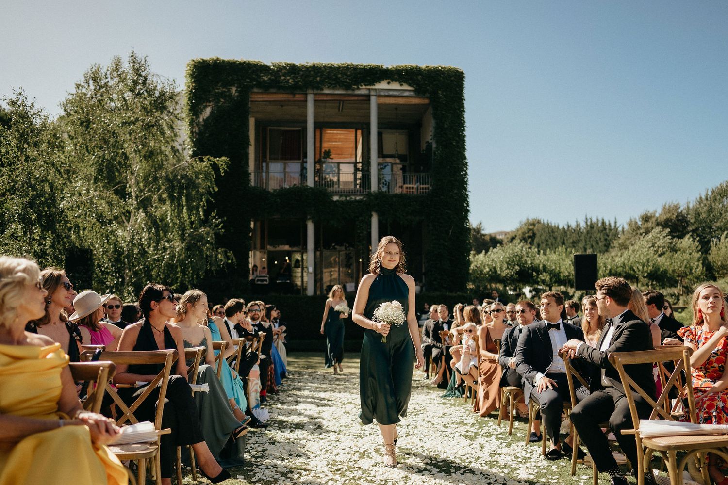 Wedding guests seated outdoors in white chairs watch ceremony under bright sunlight.