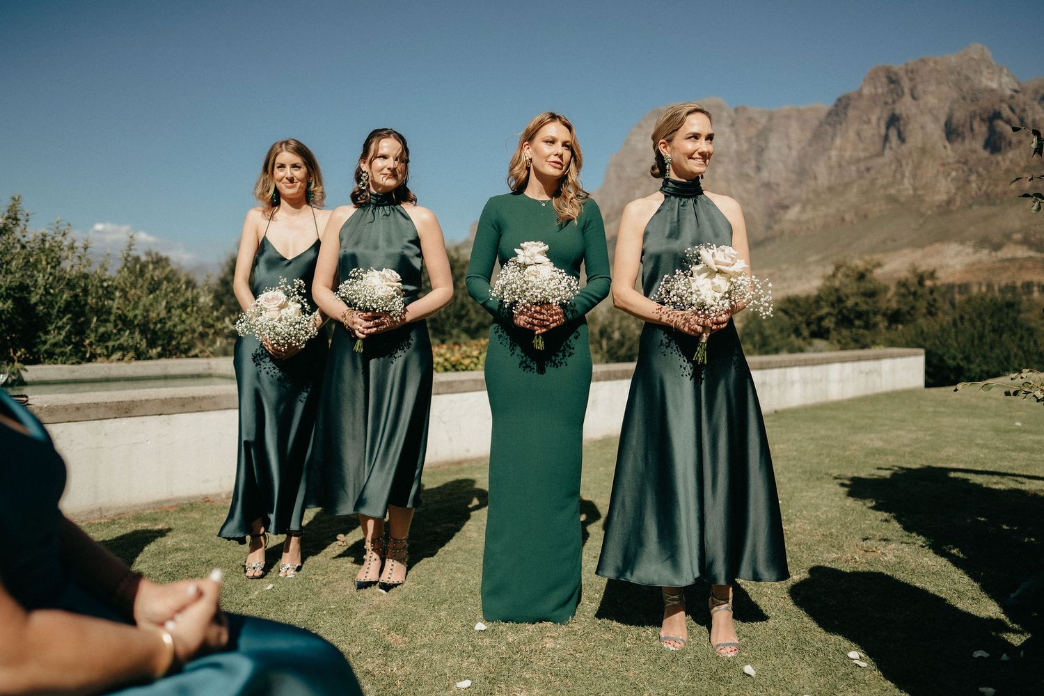 Group in emerald green bridesmaid dresses pose together against mountain backdrop on sunny day.