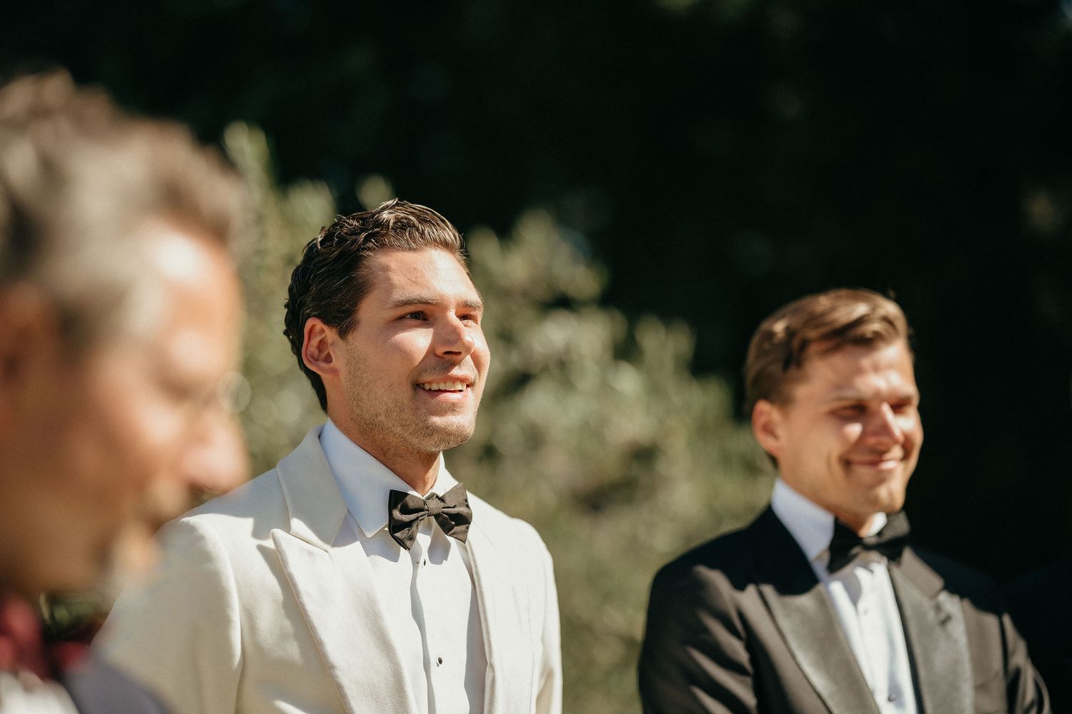 Groomsmen in black tuxedos and white bowties stand together outdoors during a wedding ceremony.