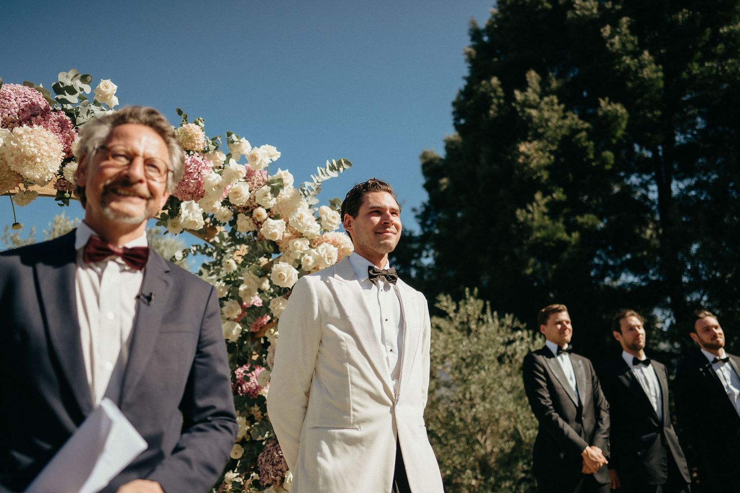 Wedding party reacts with joy and laughter during an outdoor ceremony with white floral arrangements.