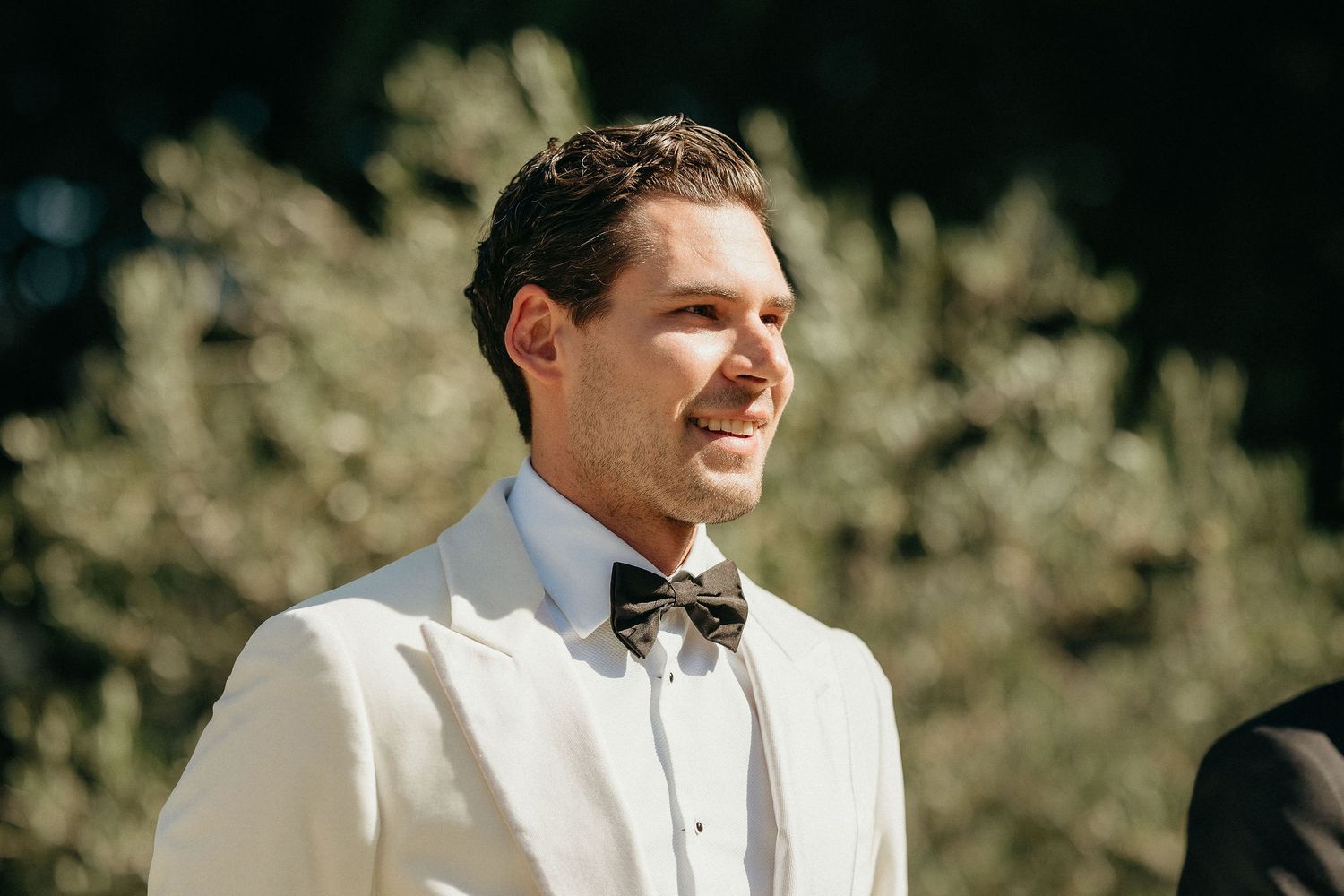 A groom in a white tuxedo jacket and black bowtie smiles warmly during an outdoor wedding ceremony.