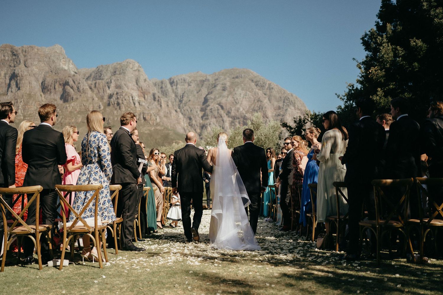 A panoramic wedding ceremony takes place against dramatic mountain cliffs in a scenic outdoor setting.