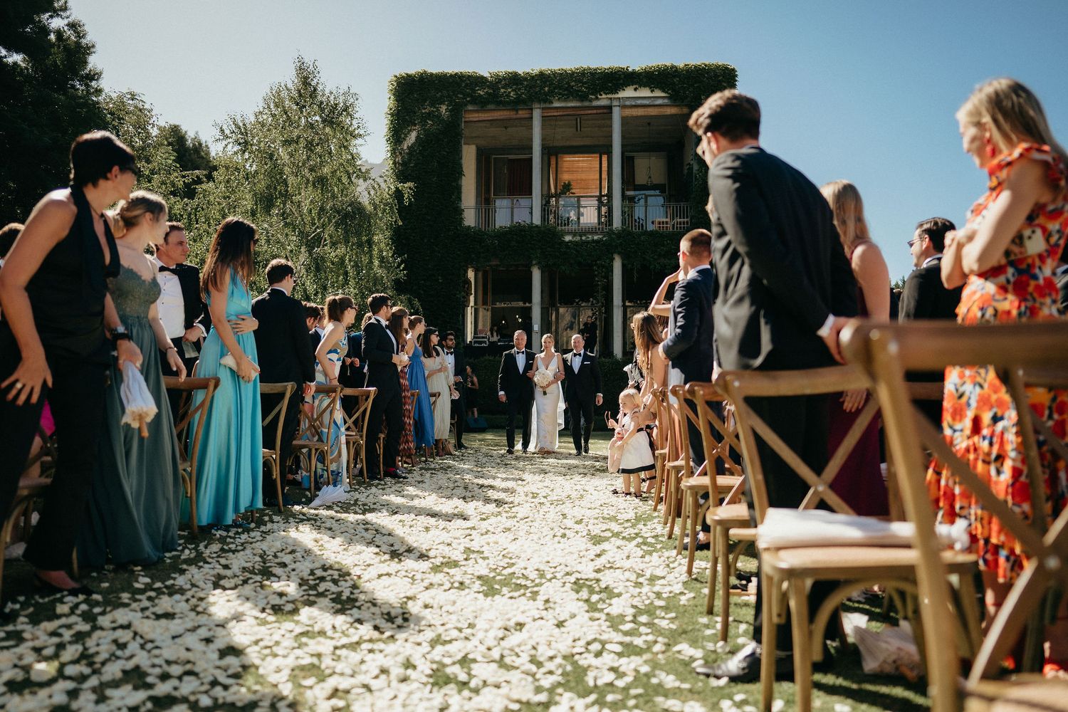 Guests stand along a petal-strewn aisle during an outdoor wedding ceremony on a sunny day.
