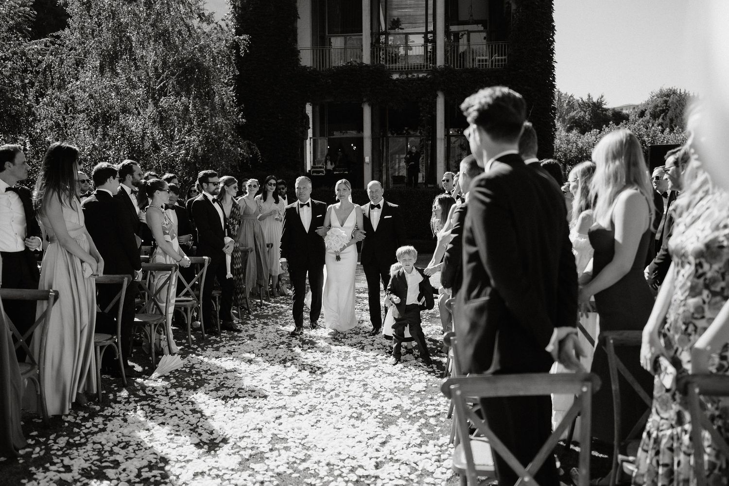 Black and white wedding ceremony photo showing guests lining a path covered in flower petals as couple walks down aisle.